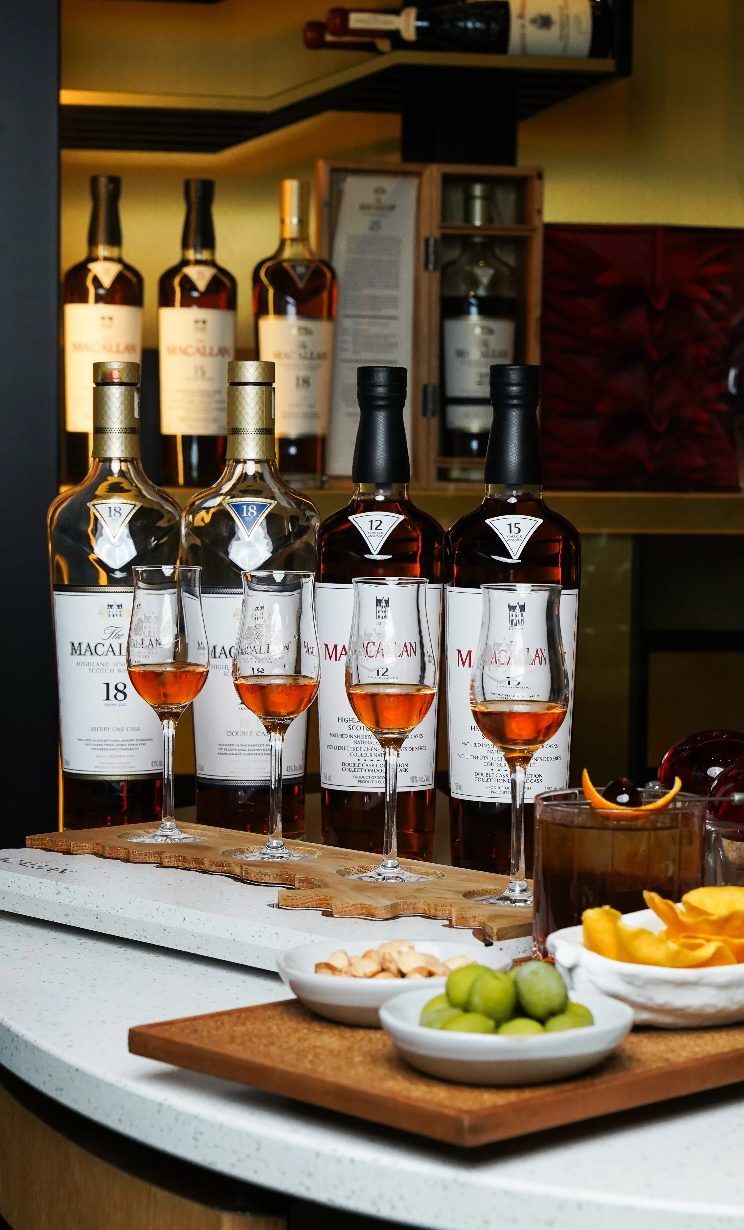 Several bottles of Macallan whiskey with glasses filled with whiskey on a wooden tray, surrounded by bowls of green grapes, nuts, and dried fruits, in a bar or tasting room setting.