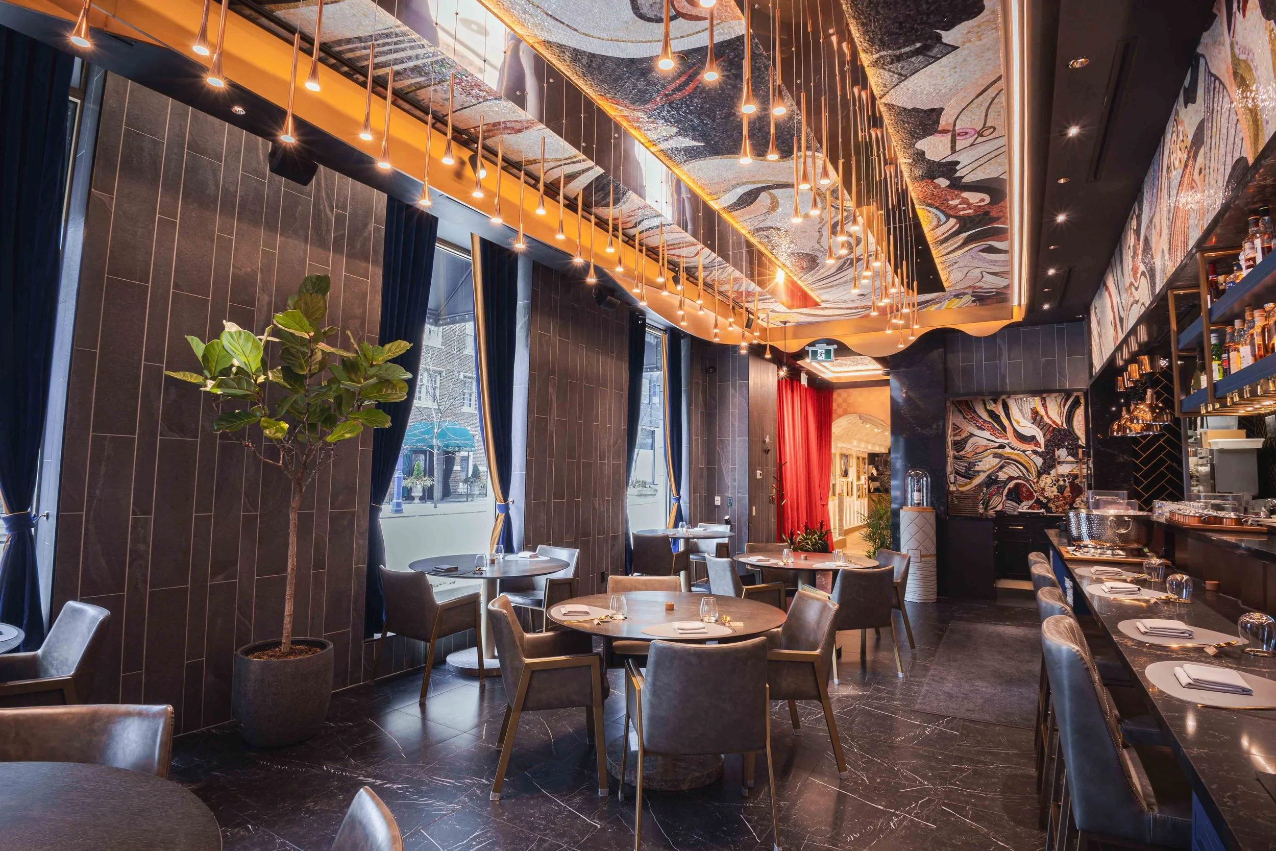 Interior of a modern restaurant with round tables, gray chairs, and a large potted plant near the window. Decor includes dark tiles, curtain panels, and artistic wall and ceiling designs with hanging lights.