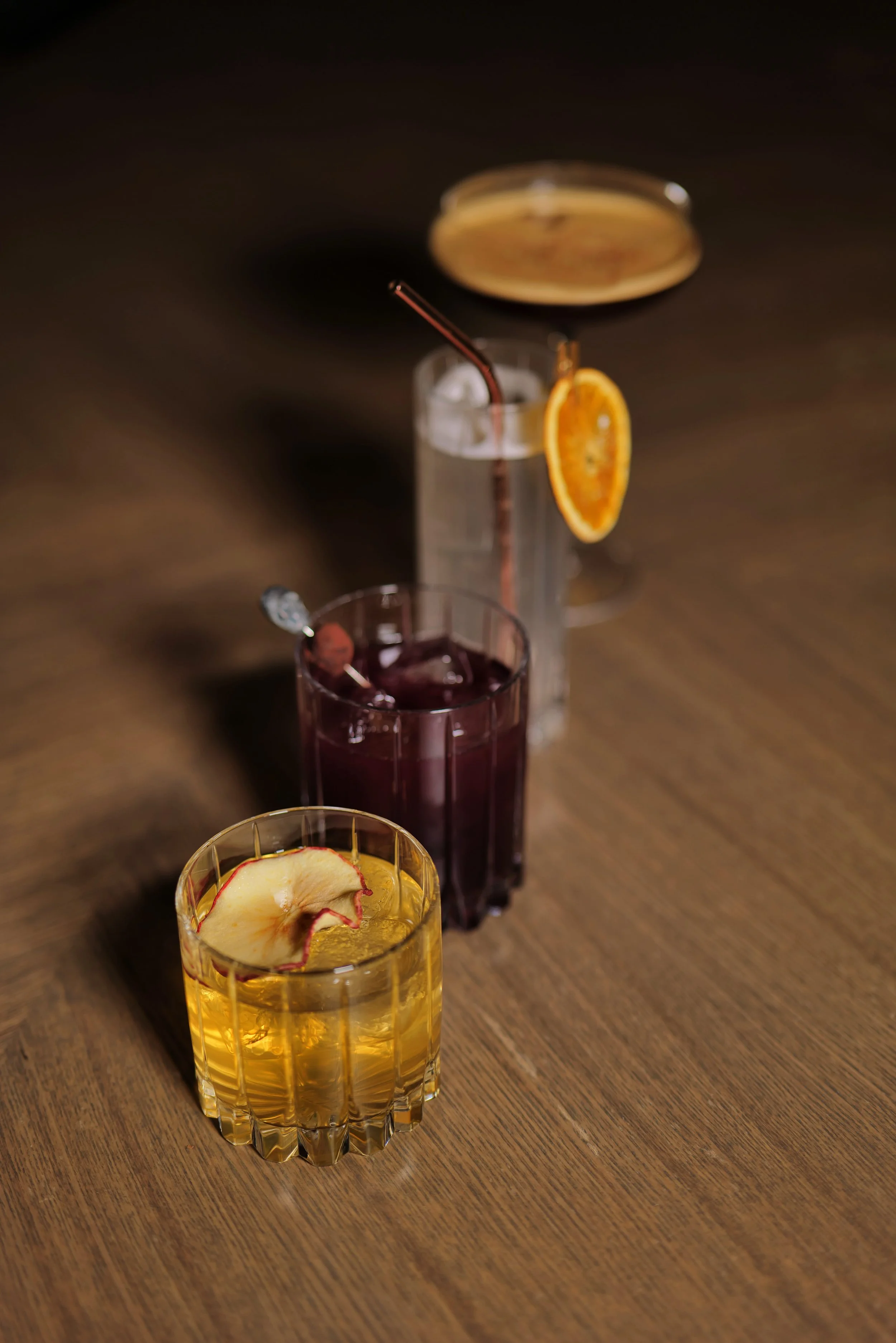 Three drinks on a wooden table; one garnished with a slice of apple, another with an orange slice and a straw, and the third with a cherry