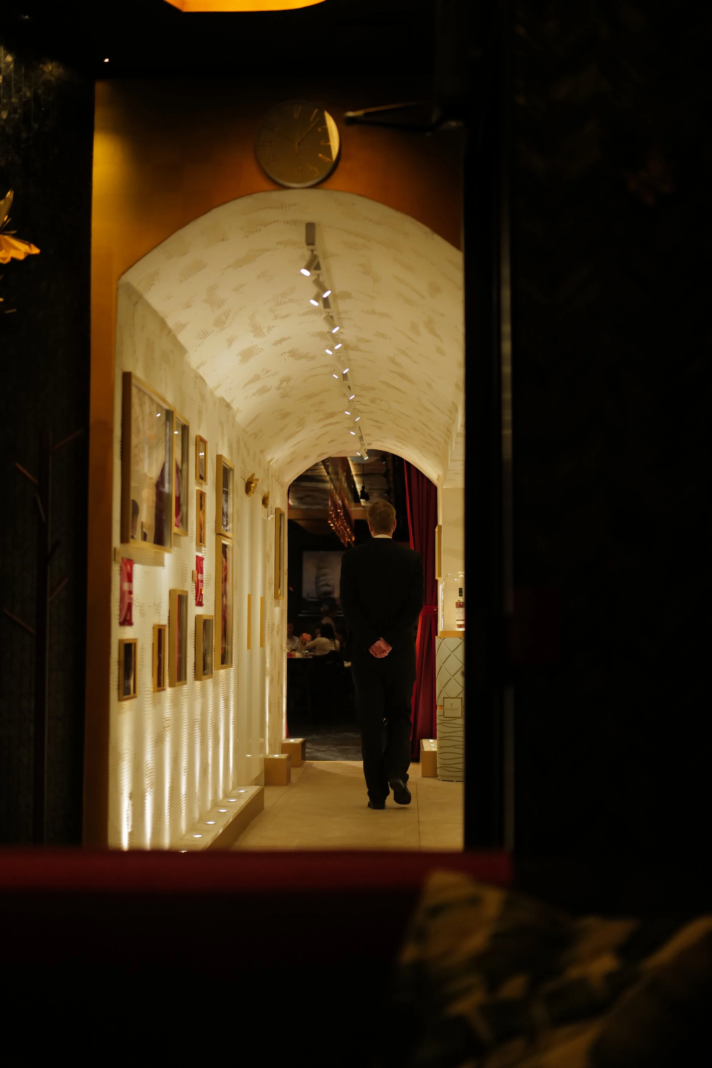 A man in a suit walking through a warmly lit hallway in a restaurant or gallery, with framed pictures on the wall and a decorative arched ceiling.