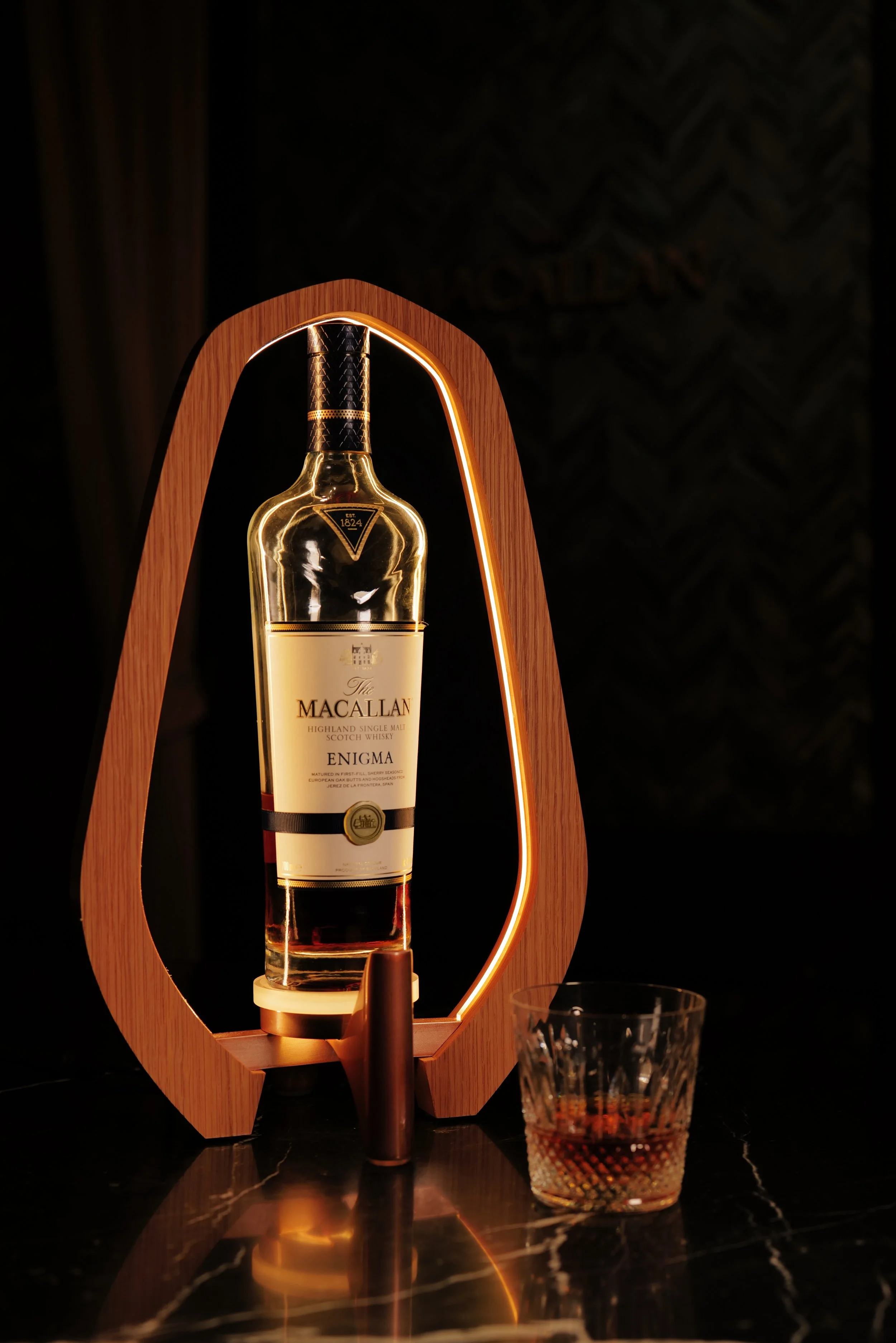 A bottle of Macallan Enigma whiskey on a wooden stand with a glowing light, and a glass of whiskey on a dark, reflective surface.