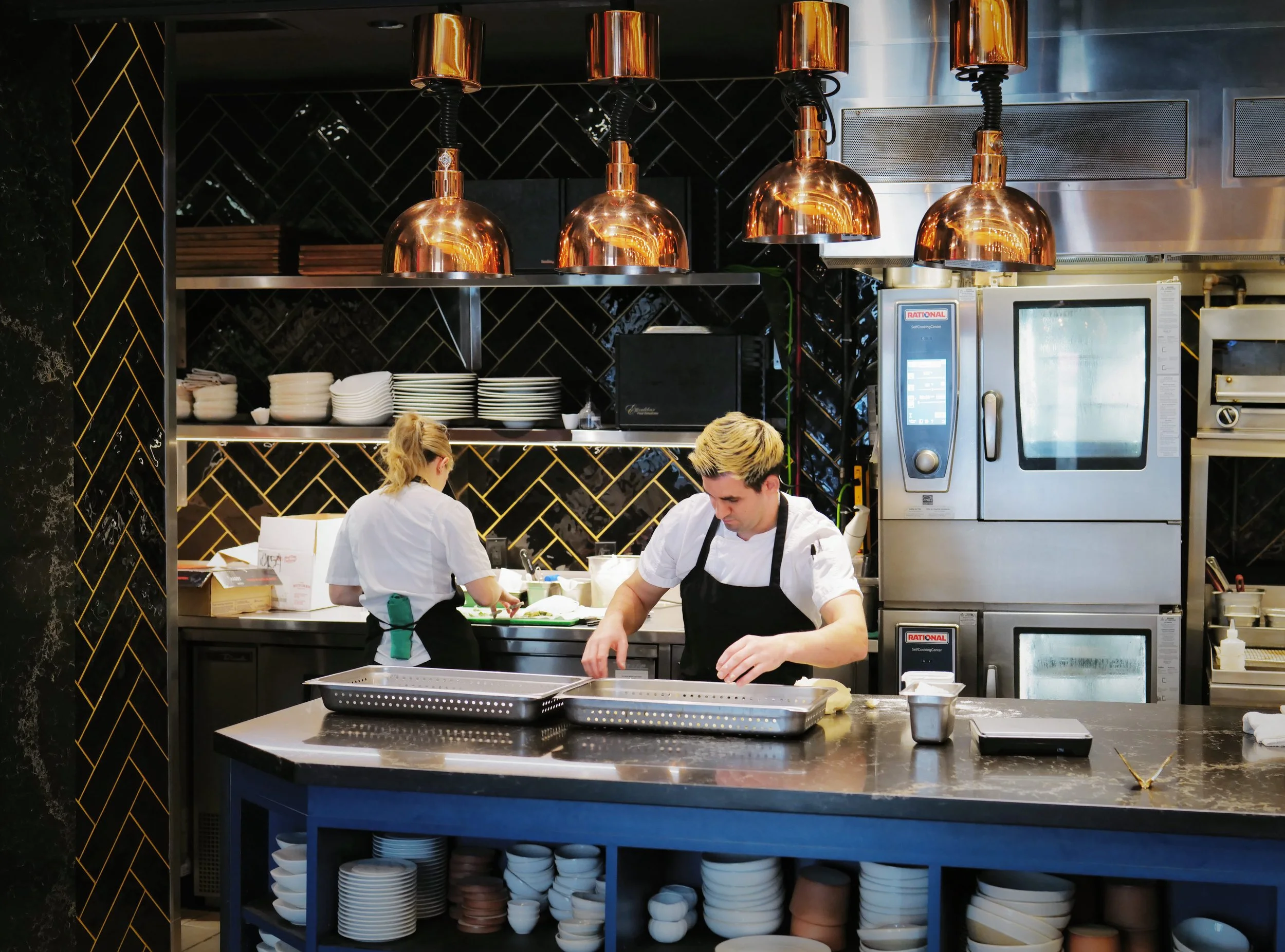 Two chefs in a professional kitchen preparing food, with one in the foreground and the other working at the back, under copper pendant lights with black and gold backsplash.