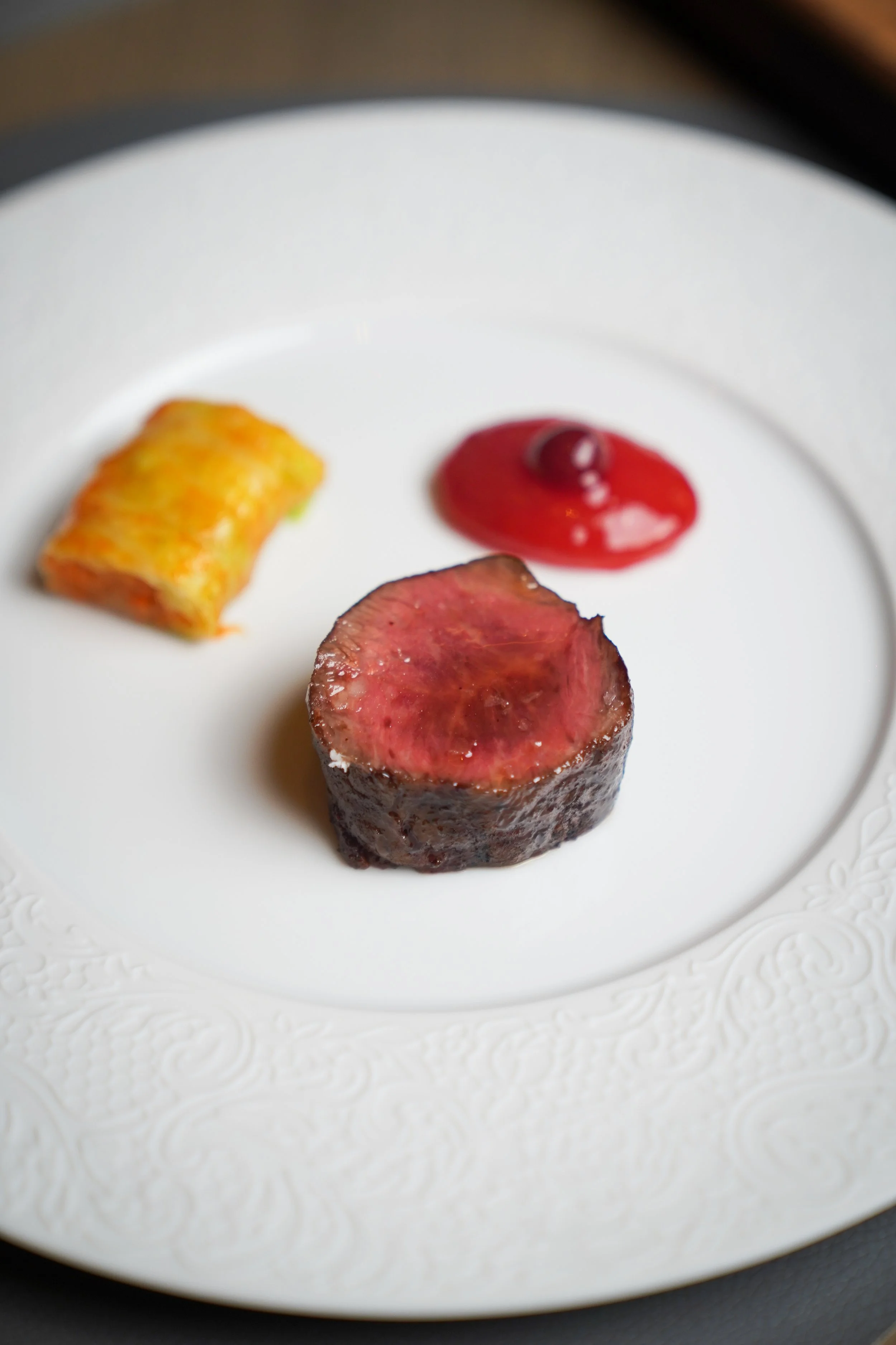 A white plate with an ornate border holding three food items: a cylindrical piece of seared beef with a pink center, a small rolled omelette, and a dollop of red sauce with a small garnish on top.