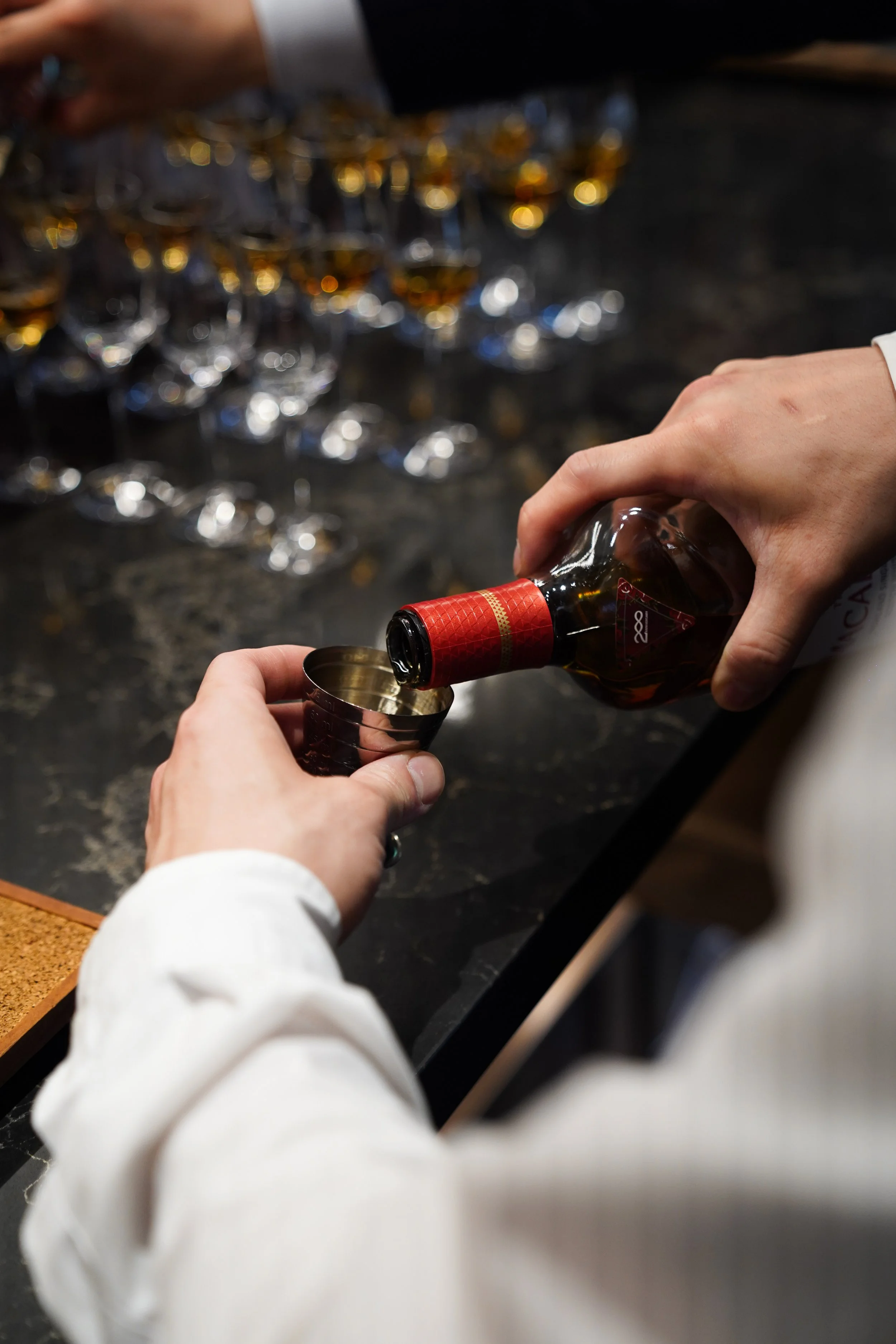 Person pouring a shot of liquor into a metal shot glass at a bar, with numerous glasses of alcohol in the background.