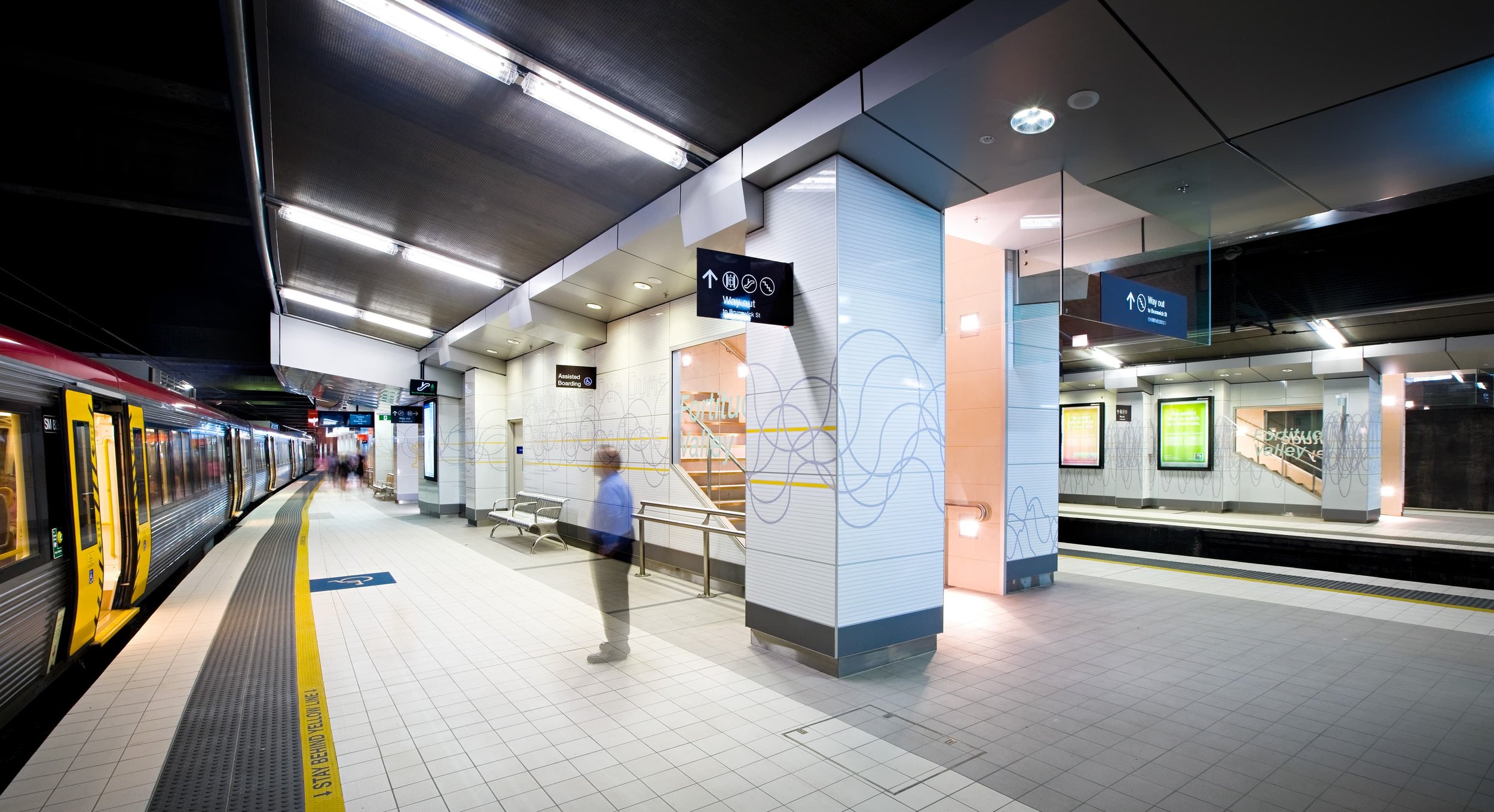 Fortitude Valley train station with a platform, a train on the left, and a few blurred people walking. Signage indicates directions for the train and amenities. Bright lighting and tiled floors are visible.