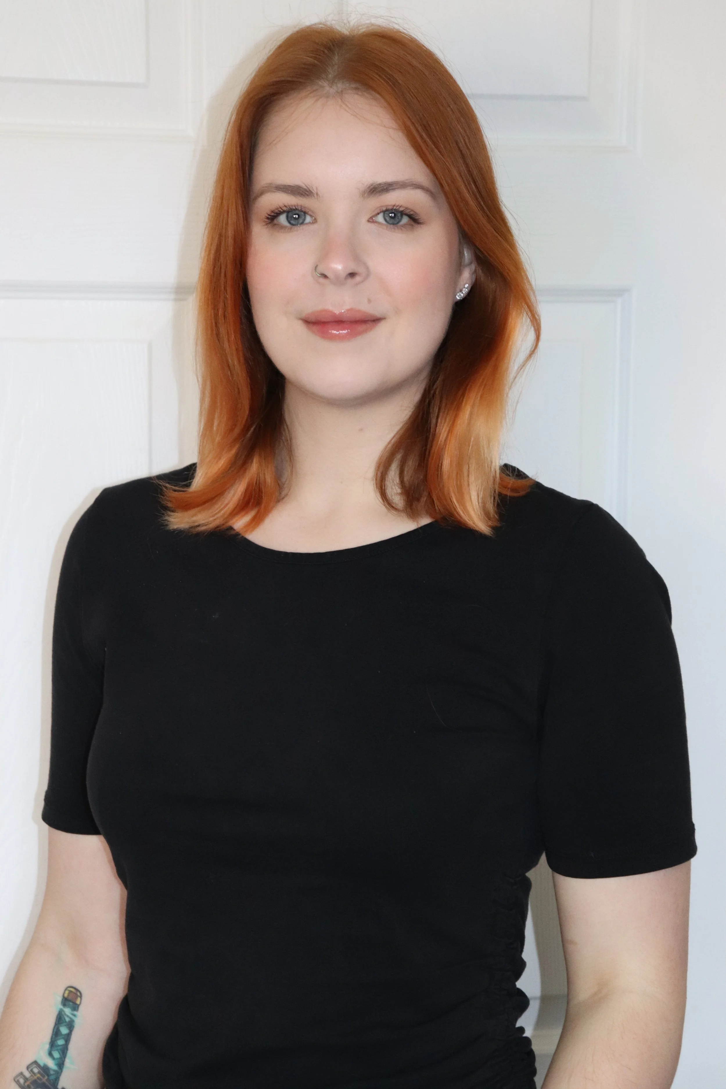 A woman with red hair and light skin, wearing a black top, standing in front of a white door, with tattoos on her left arm.