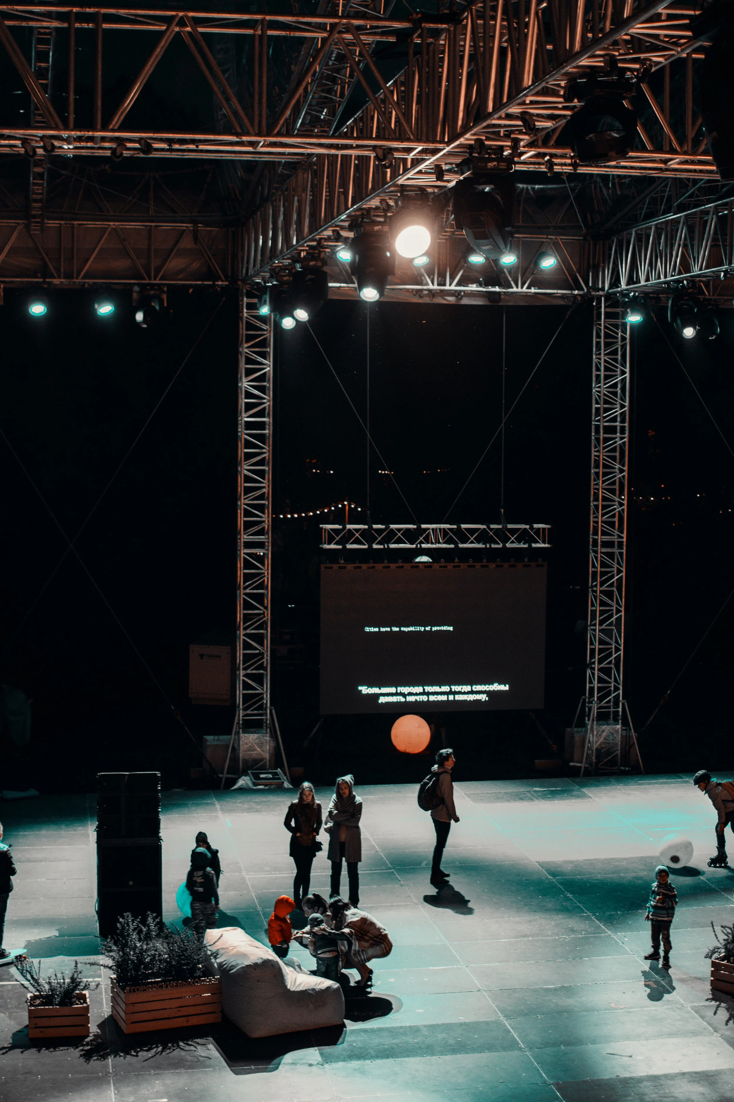 An outdoor stage setup at night with a large LED screen displaying text, surrounded by metal scaffolding with bright stage lights. There are a few people, including children, on the ground near some planters and seating, indicating a casual or concert event.