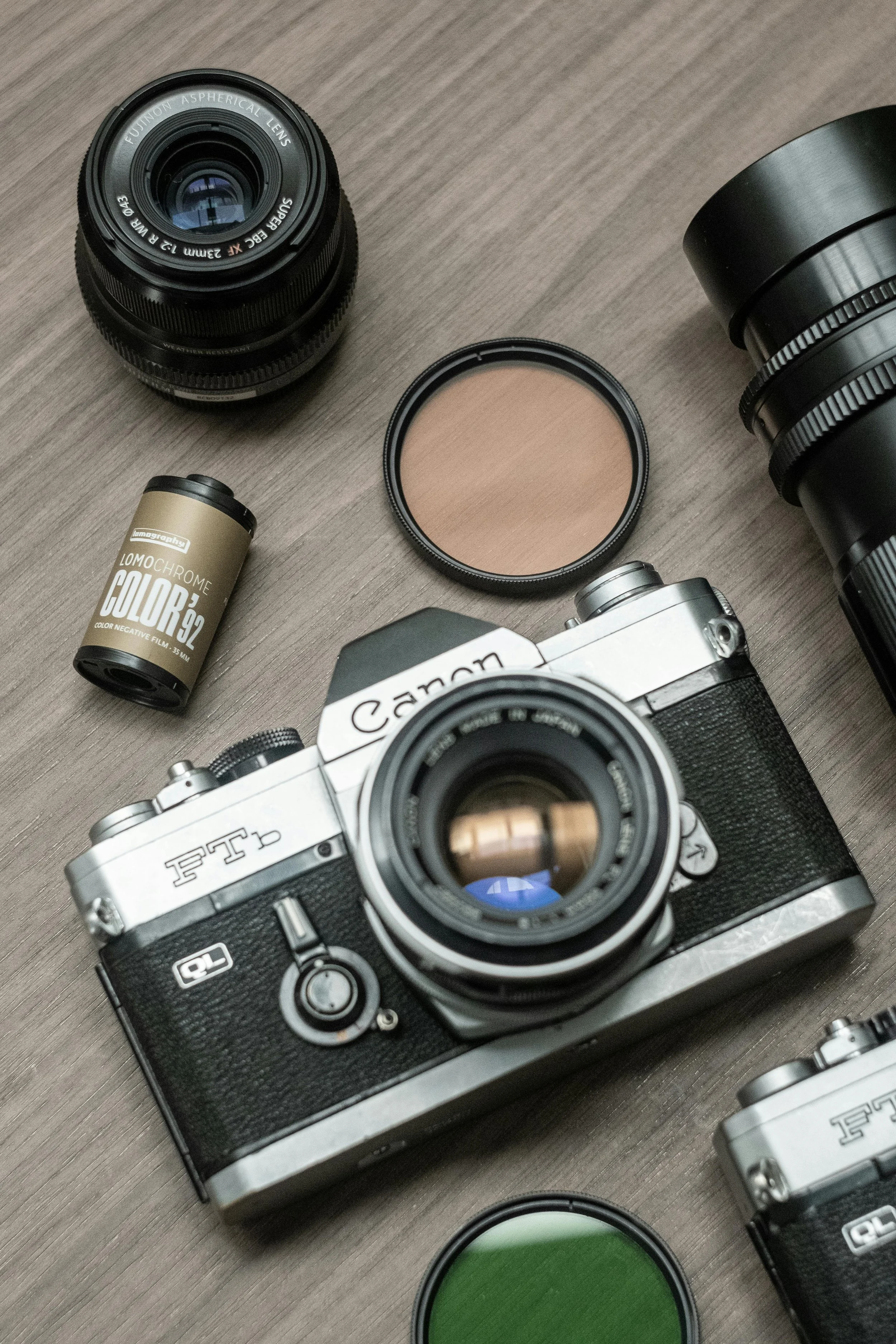 A collection of vintage and modern camera equipment, including a Canon film camera, camera lenses, a roll of Lomochrome color film, and color filters, arranged on a wooden surface.