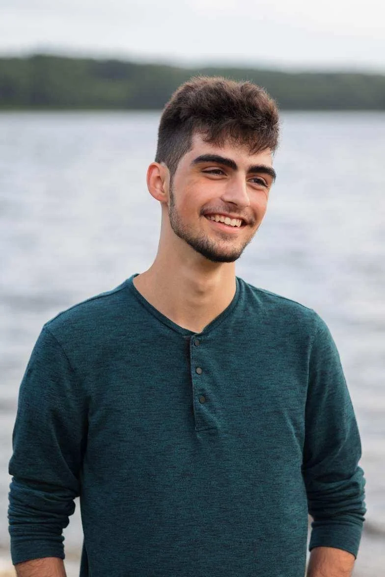 A young man with dark hair, facial hair, and a smile, standing outdoors near a body of water with trees in the background.