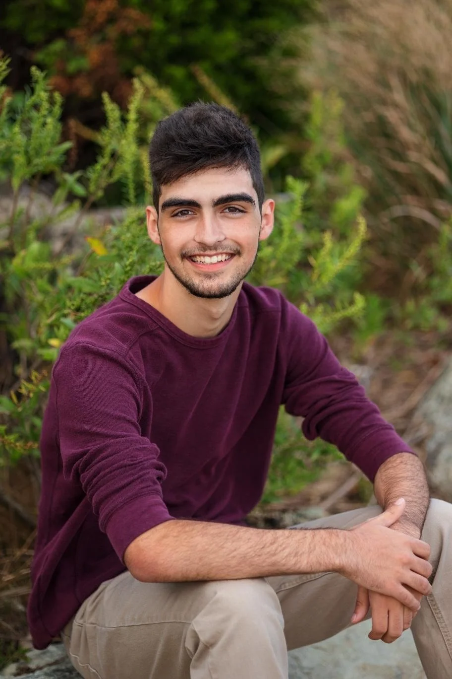 Young man with dark hair and a beard smiling, wearing a maroon long-sleeved shirt, sitting outdoors on rocks with greenery in the background.