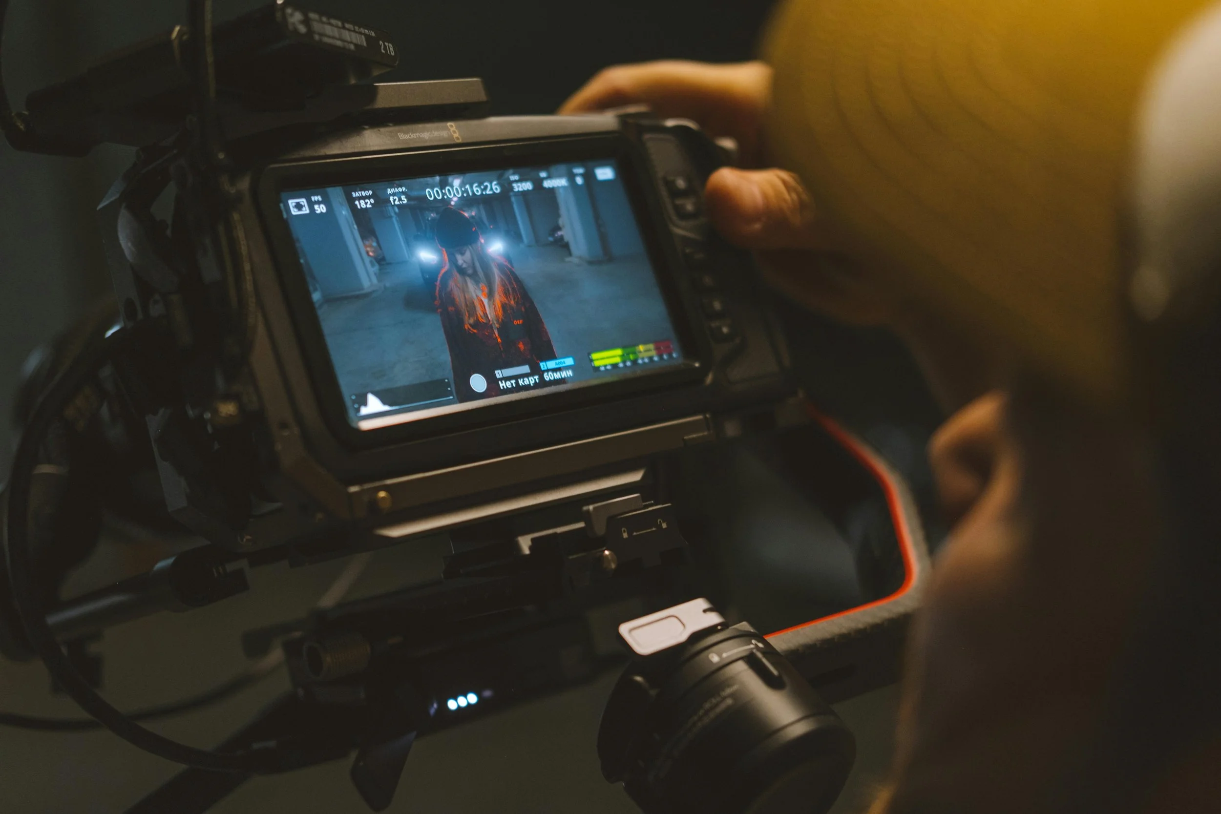 A person filming a scene with a professional video camera in an indoor parking garage. The camera's LCD screen shows a woman wearing a hat and a jacket with bright lights behind her.