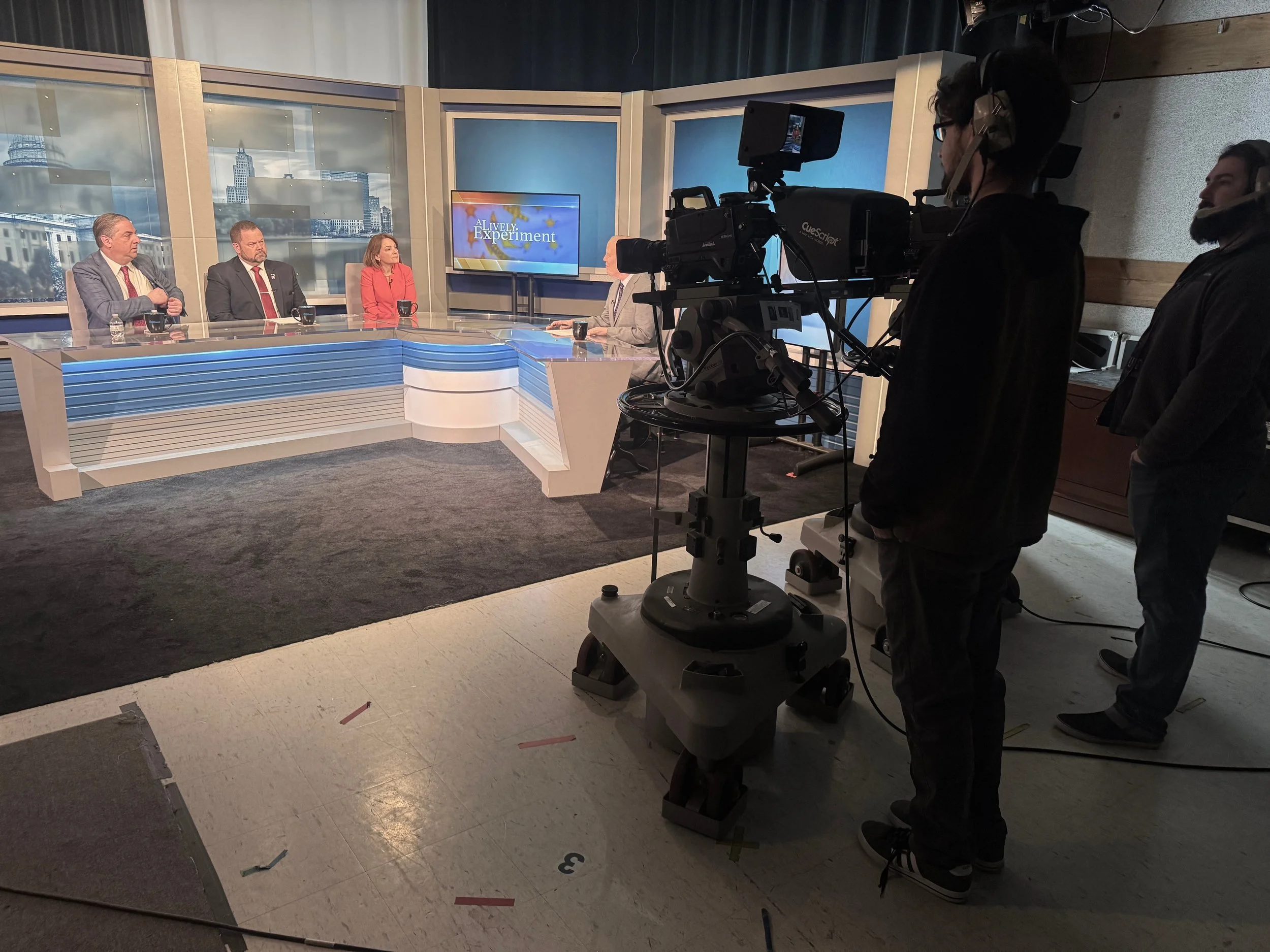 Television studio with four people seated at a news desk, a host interviewing them, camera operators filming the segment, and a large screen in the background displaying the show's title.