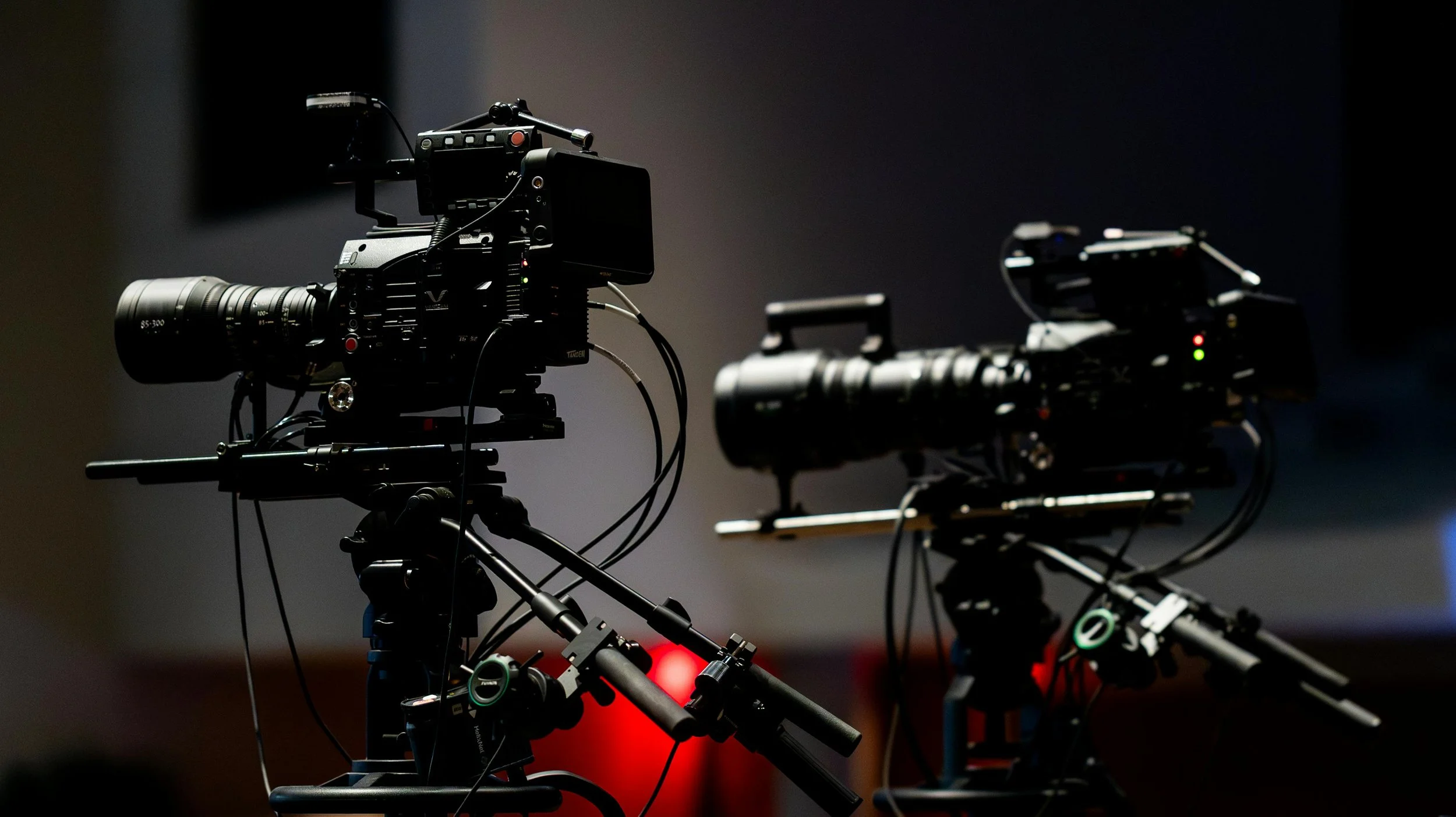 Two professional video cameras on tripods in a darkened studio or control room.