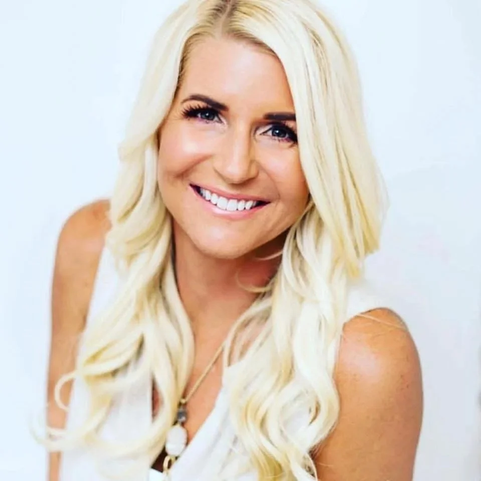 A smiling woman with long, wavy blonde hair, wearing a white sleeveless top, standing against a plain white background.
