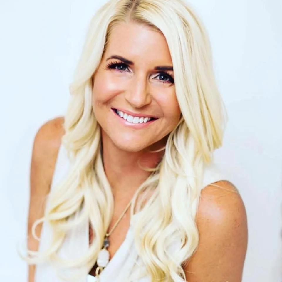 A smiling woman with long, wavy blonde hair and blue eyes, wearing a white sleeveless top and a necklace with a pendant, standing against a plain white background.