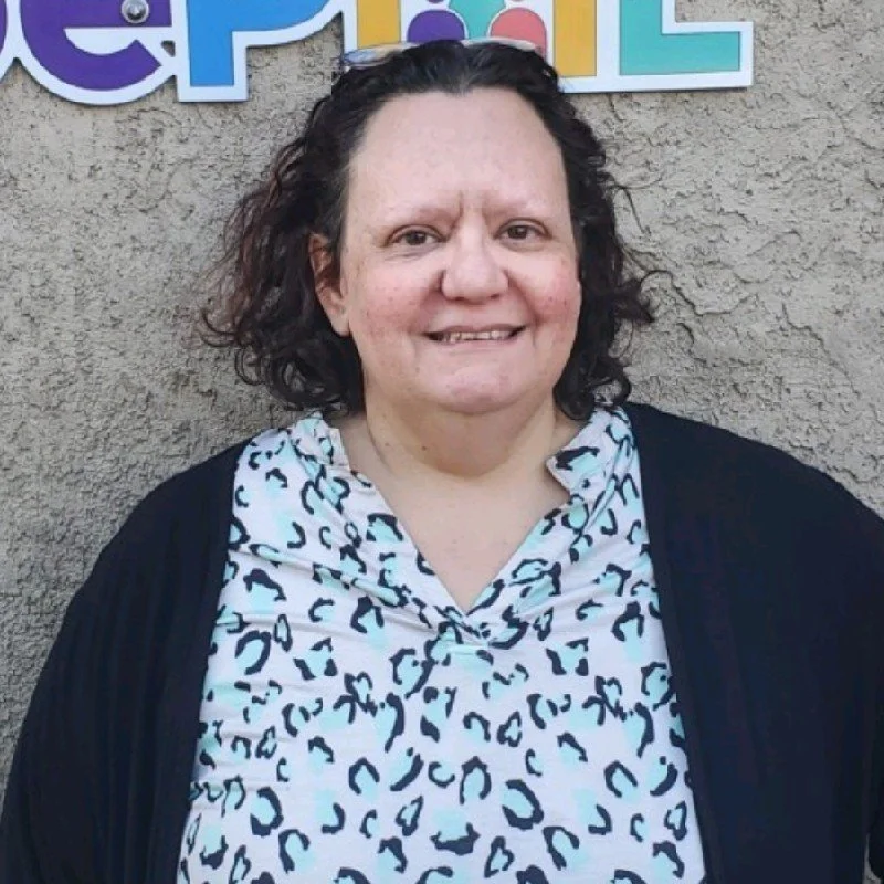 A woman with shoulder-length curly hair, wearing a black cardigan over a white leopard-print shirt, standing in front of a textured gray wall with colorful letters and shapes in the background.