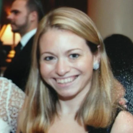 A smiling woman with blonde hair at a social event, with a blurred man in a suit in the background.