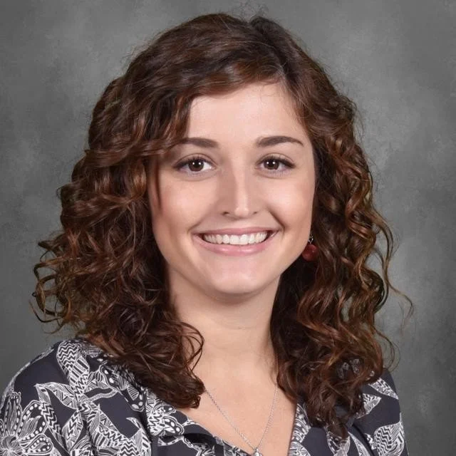 A young woman with curly brown hair, smiling, wearing a patterned black and white top and cherry earrings, against a gray background.