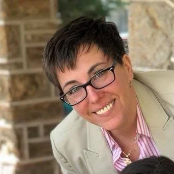 A woman with short dark hair, glasses, and a light-colored blazer smiling in an outdoor setting with stone and brick background.