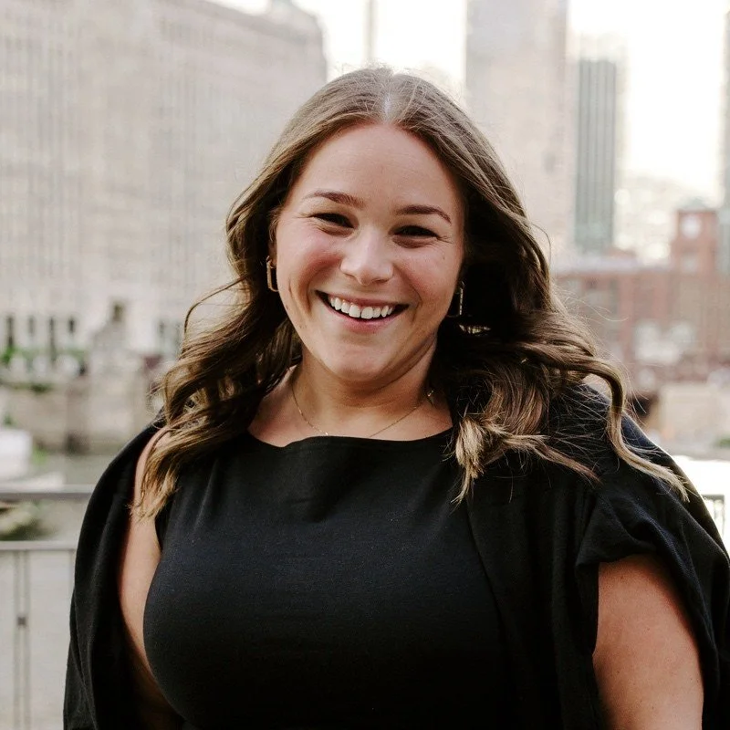 Smiling woman with wavy brown hair, wearing a black top, standing outdoors with city buildings in the background.