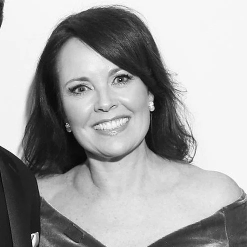 A woman with shoulder-length dark hair, wearing earrings and smiling, dressed in a shiny off-the-shoulder top.