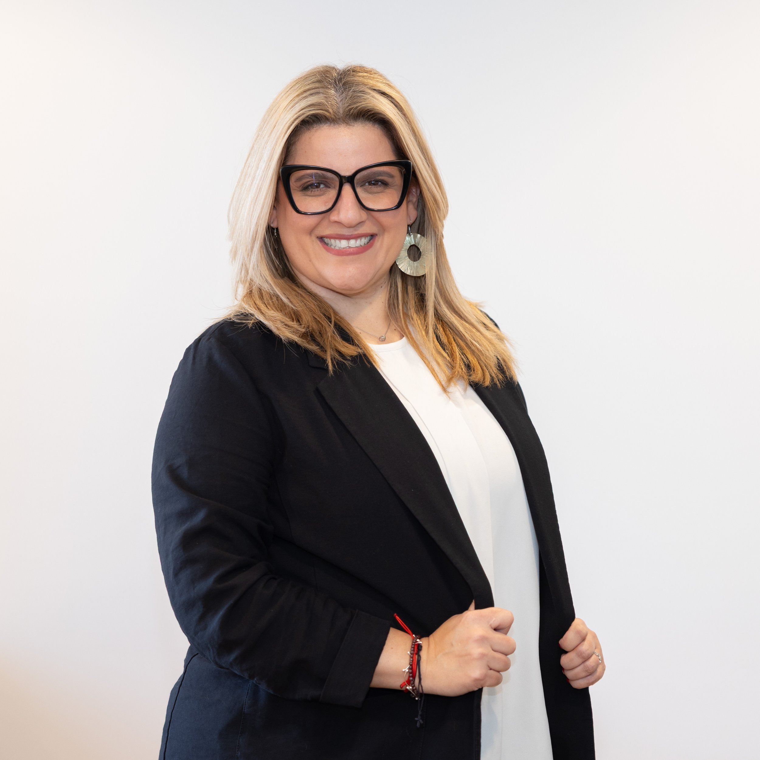 A woman with blonde hair wearing glasses, a black blazer, and a white top, smiling and standing against a plain white background.