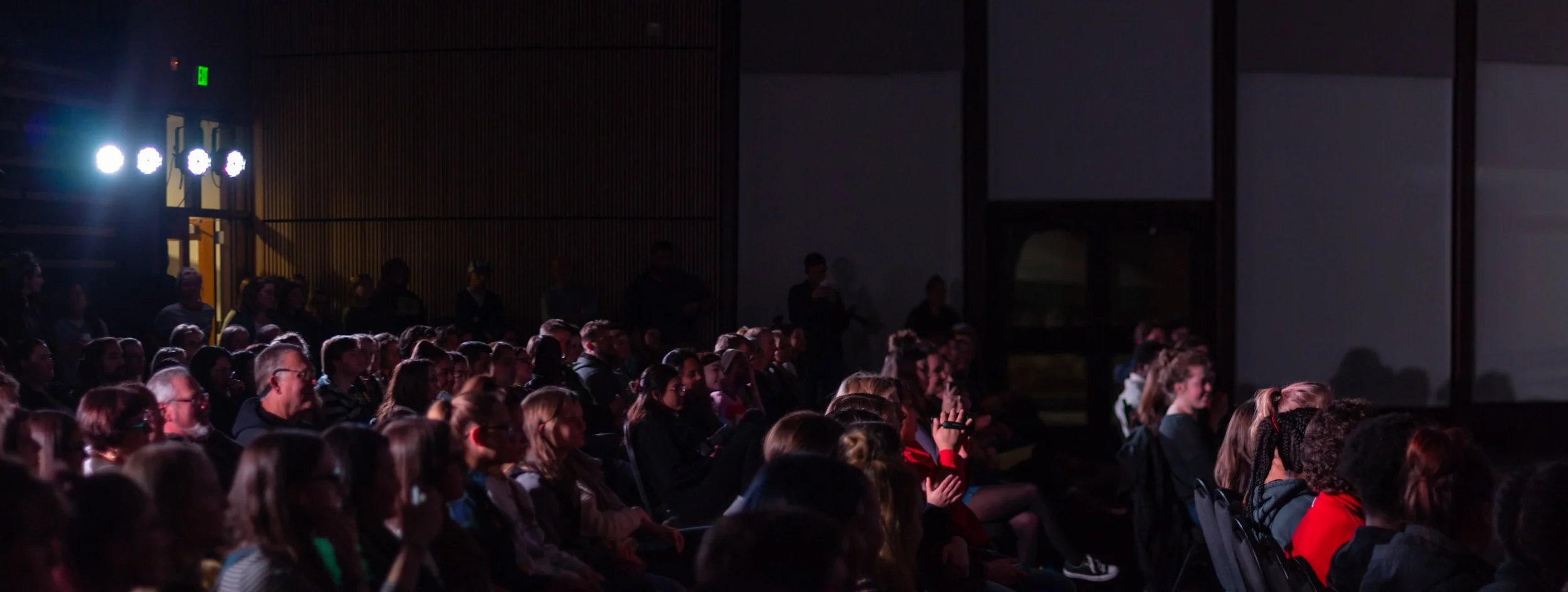 Audience seated in a dark theater or auditorium watching a performance or presentation, with some children and adults visible.