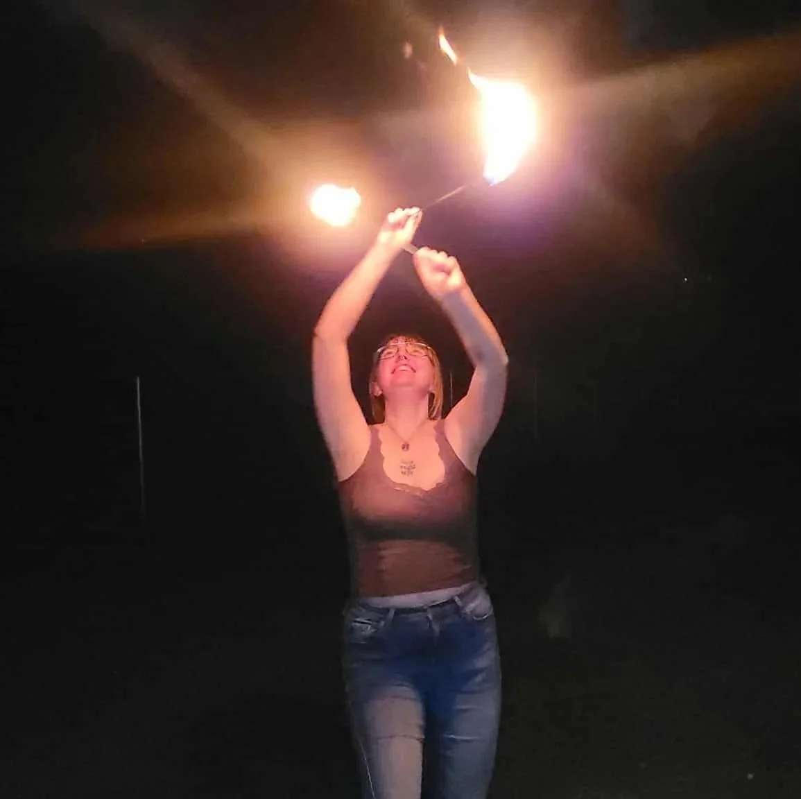 A woman holding fire wands in the dark, smiling and looking up at the flames.