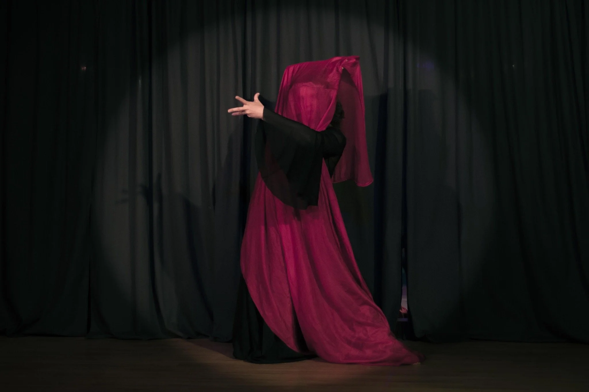 A person performing on stage, partially obscured by a pink and black veil, with black curtains in the background.