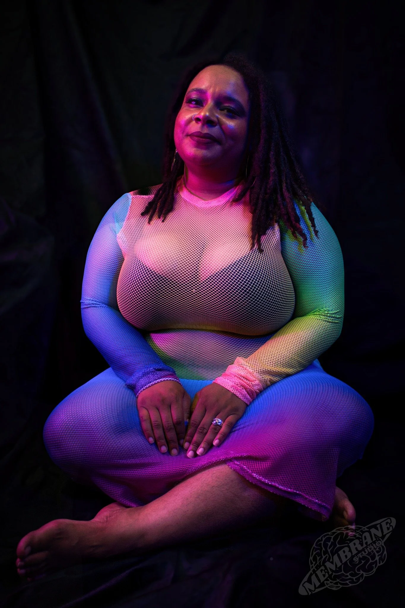 A woman with long dreadlocks wearing a multicolored mesh outfit, sitting cross-legged against a dark background, illuminated with colorful lighting.
