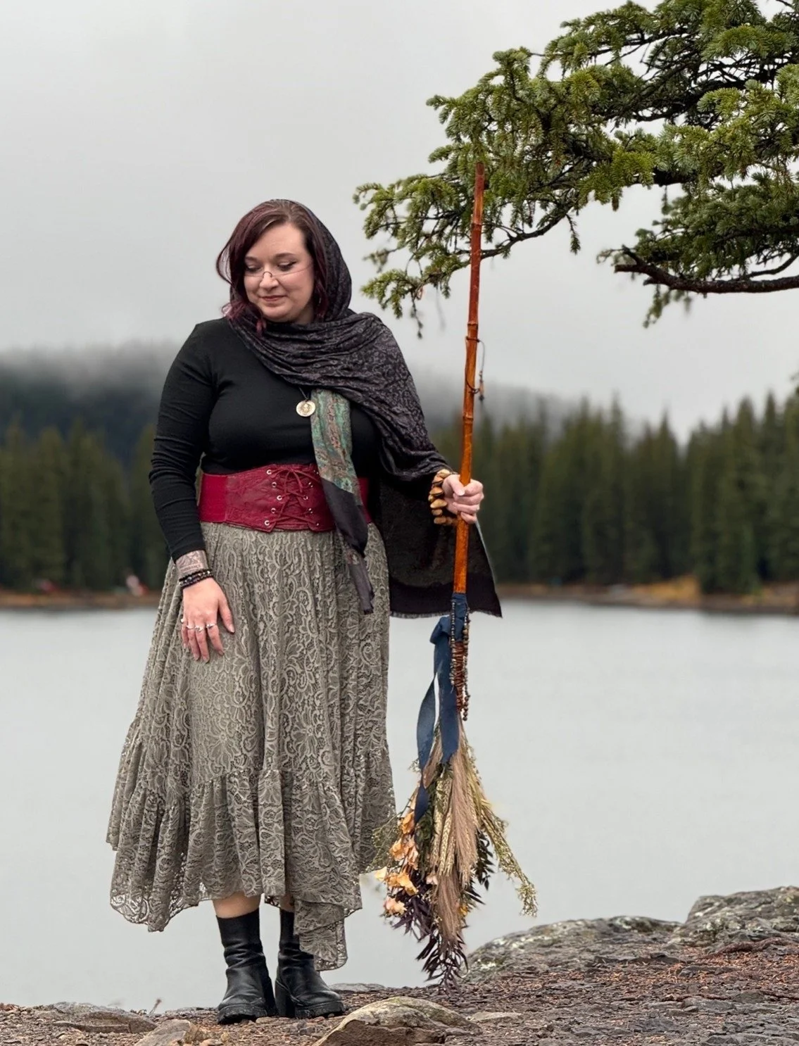 Woman outdoors near a lake, holding a staff decorated with dried floral arrangements, dressed in a patterned skirt, black top, and red waist belt, with a dark scarf over her head and shoulders, standing on rocky ground with forest in the background.
