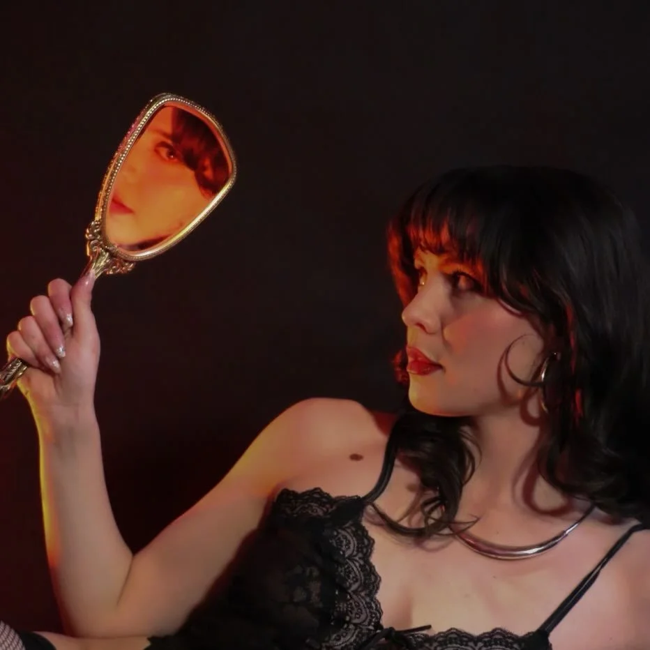 A woman with dark hair and wearing a black lace top holding a hand mirror reflecting her face.