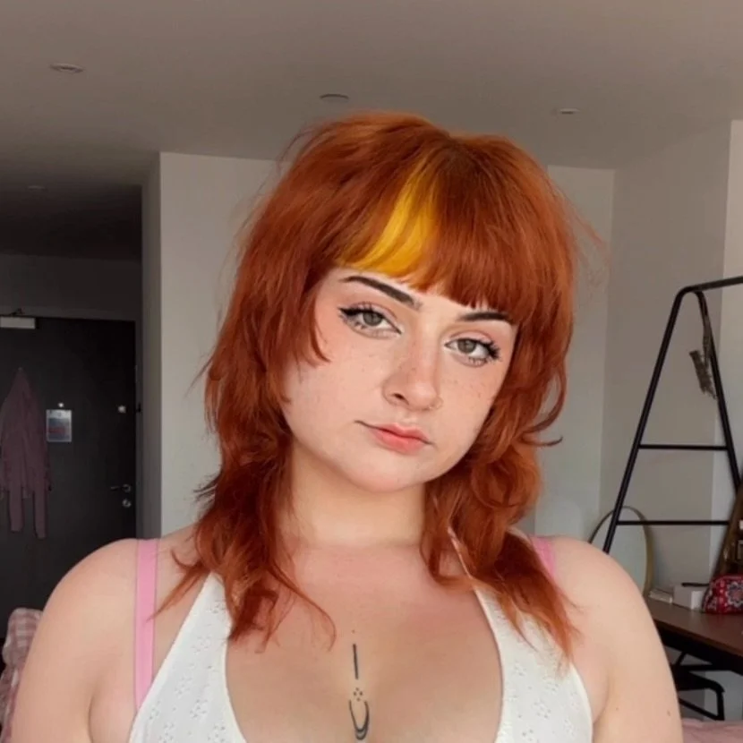 Young femme with reddish hair and a yellow streak, wearing a white top with pink straps, facing camera indoors.