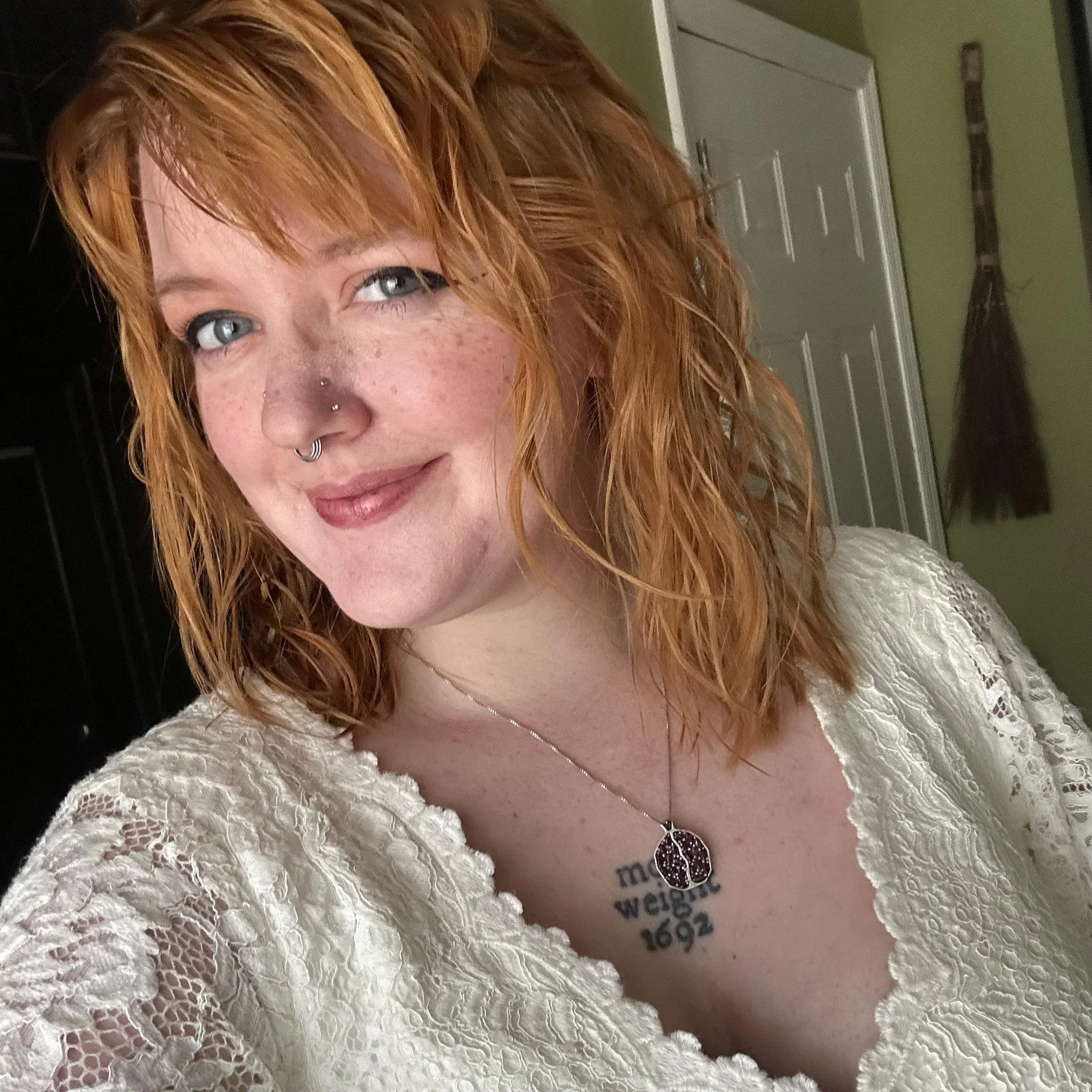 A woman with red hair, blue eyes, and freckles taking a selfie in a room. She's wearing a white lace top, a necklace with a pomegranate pendant, and has a multiple nose piercings. There is a door and hanging broom in the background.