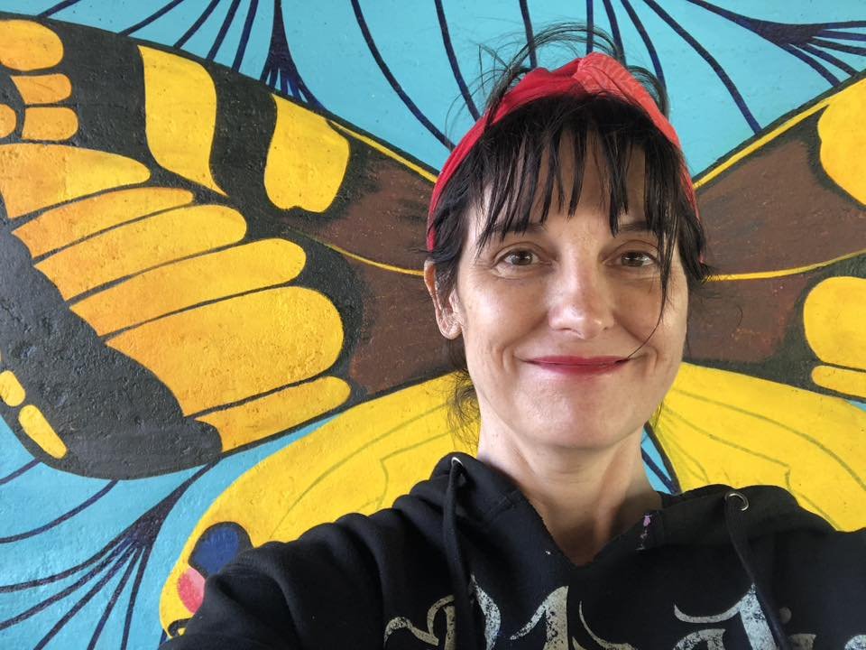 A woman with dark hair, wearing a red headband, smiling in front of a colorful butterfly mural.