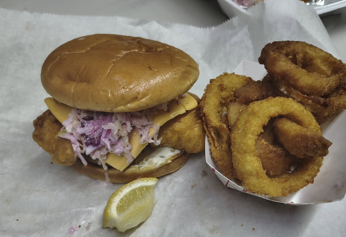 Fried fish sandwich with coleslaw and cheddar cheese in a bun, served with a lemon wedge, next to a basket of fried onion rings.