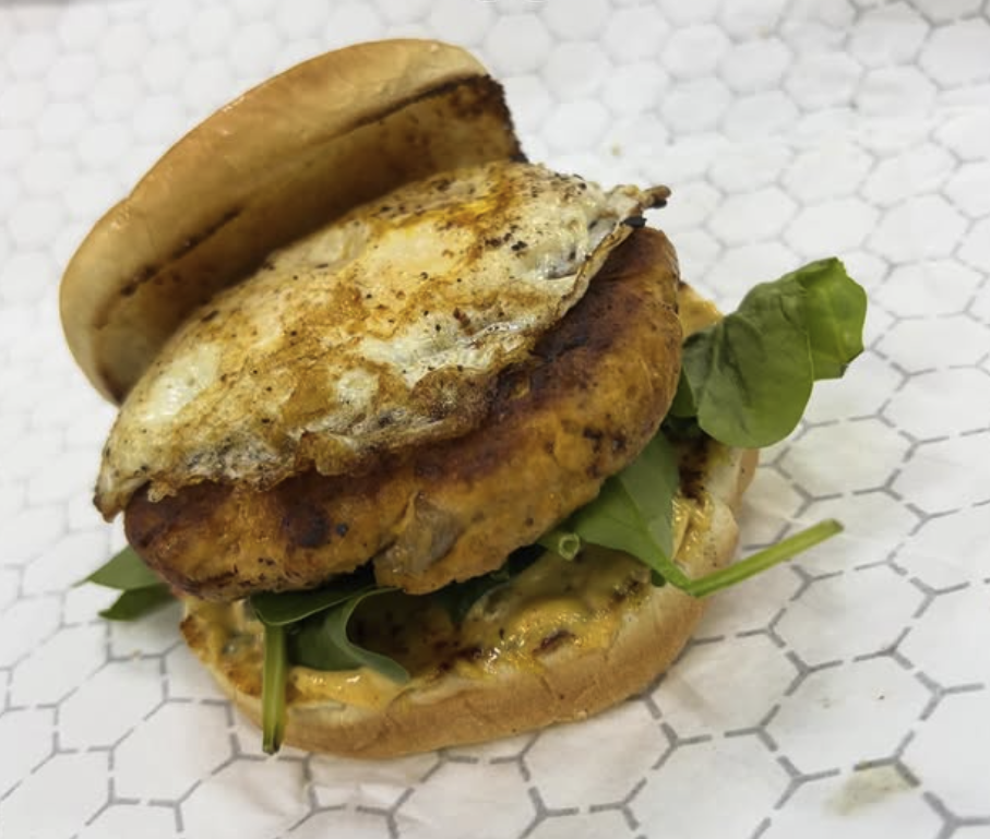 Fried egg, grilled chicken fillet, fresh greens, and sauce on a sandwich bun, served on white patterned paper.