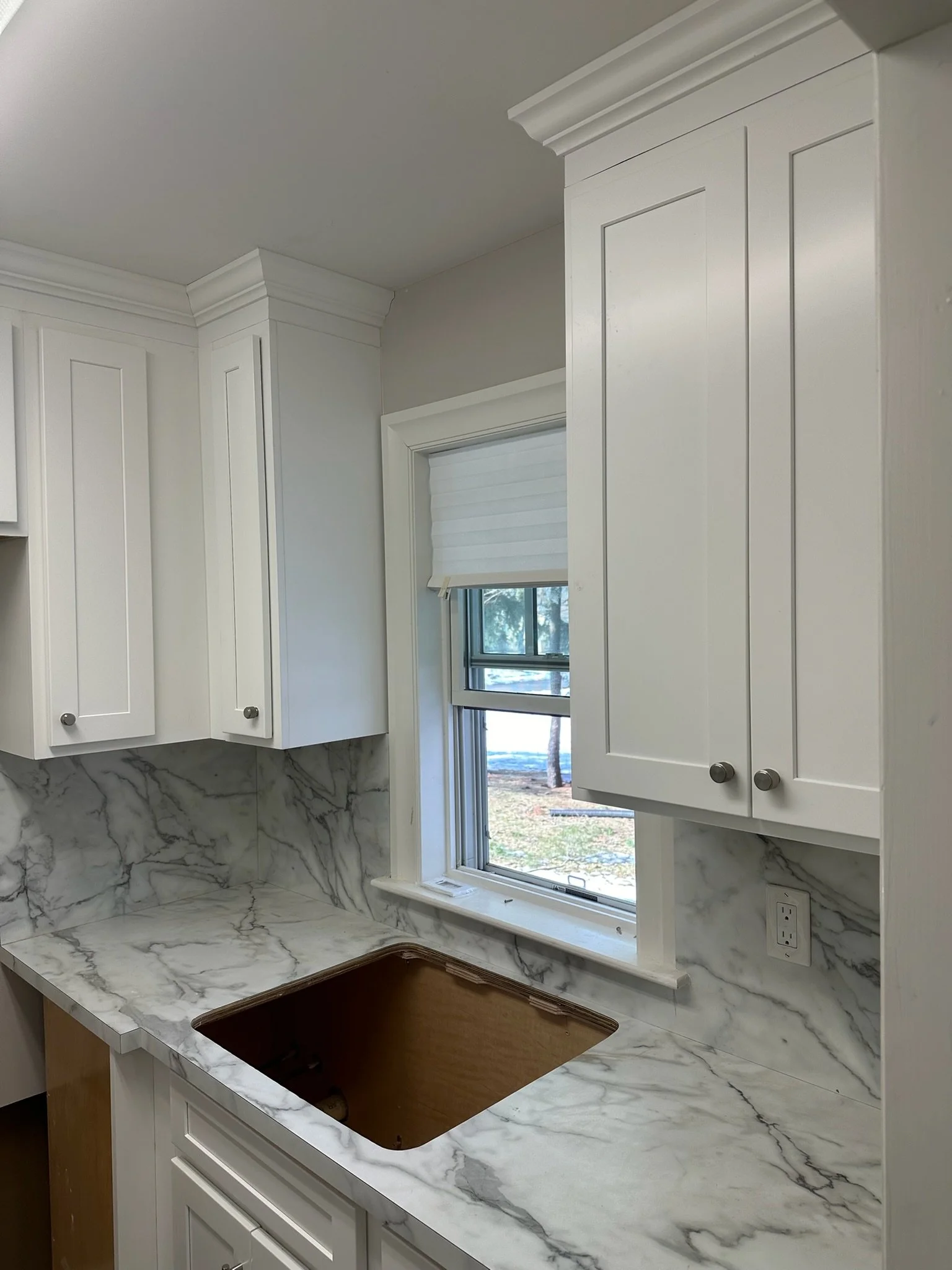 Kitchen with white cabinets, marble countertop and backsplash, a window with a white window shade, and a cutout for a sink in the countertop.