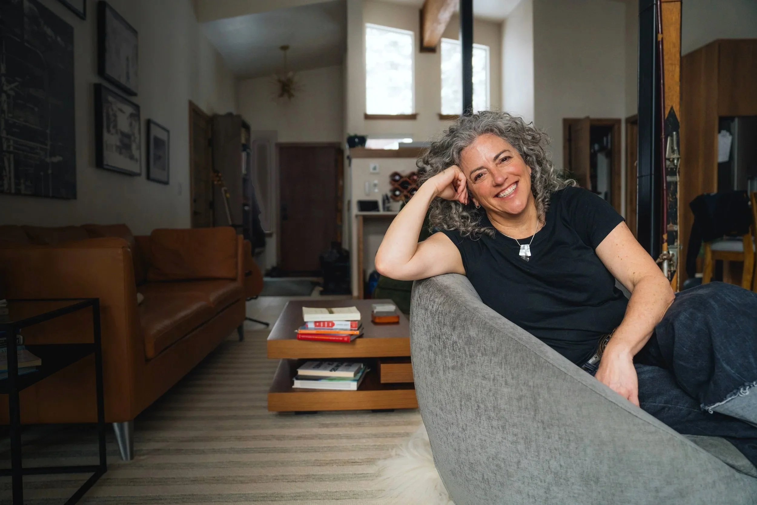 A smiling woman with curly gray hair sitting on a gray couch in a cozy living room, with a wooden coffee table and books in front of her, and a background of large windows, artwork on the walls, and wooden beams.