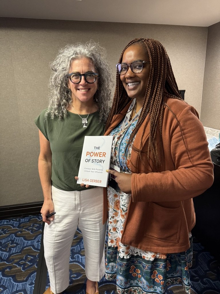 Two women standing side by side indoors, smiling at the camera. One woman has curly gray hair, glasses, and is wearing a green top with white pants. The other woman has long braided hair, glasses, and is wearing a brown jacket over a patterned dress. The woman with curly hair is holding a book titled 'The Power of Story' by Lisa Gerber.