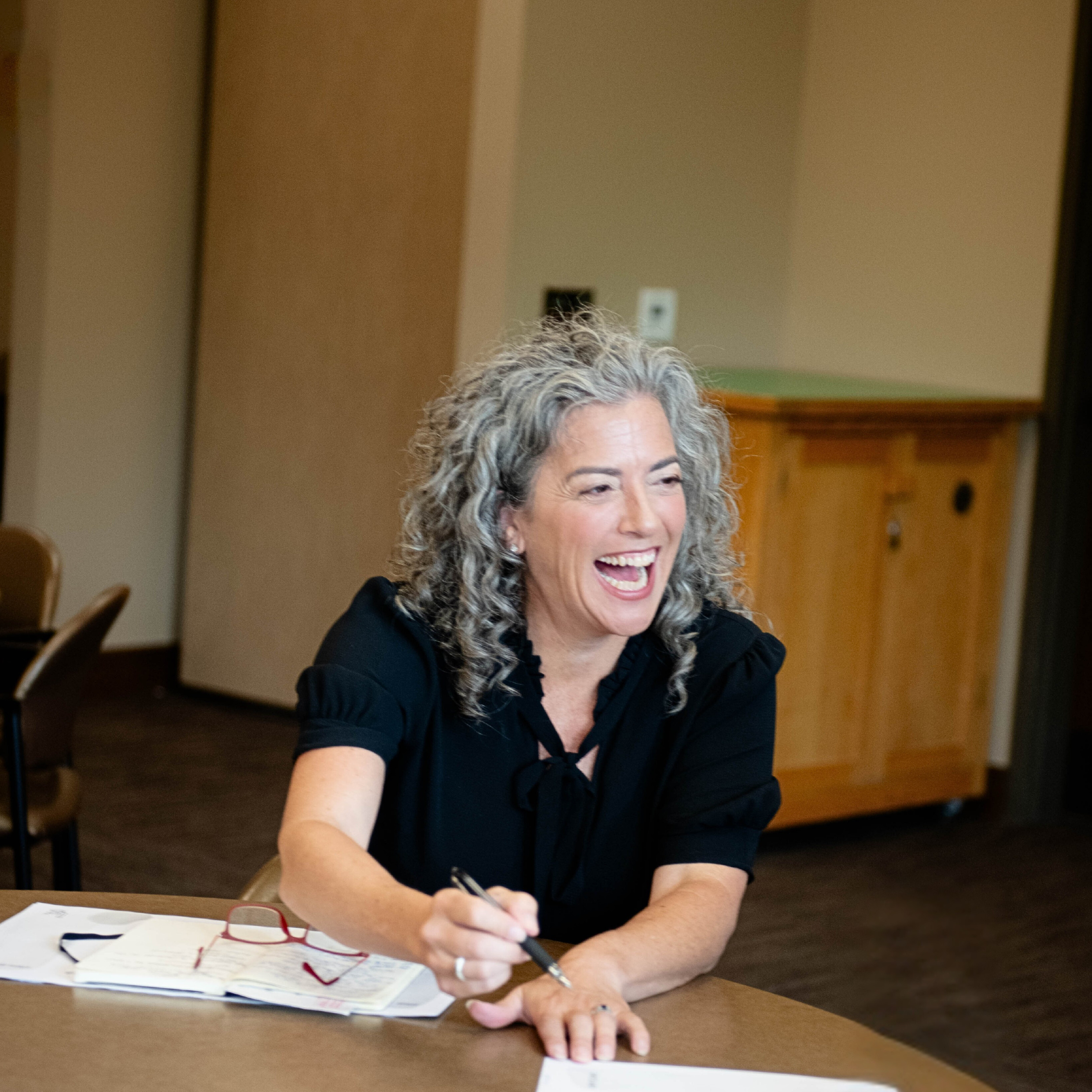 A woman with curly gray hair wearing a black top sits at a table, smiling and writing in a notebook.