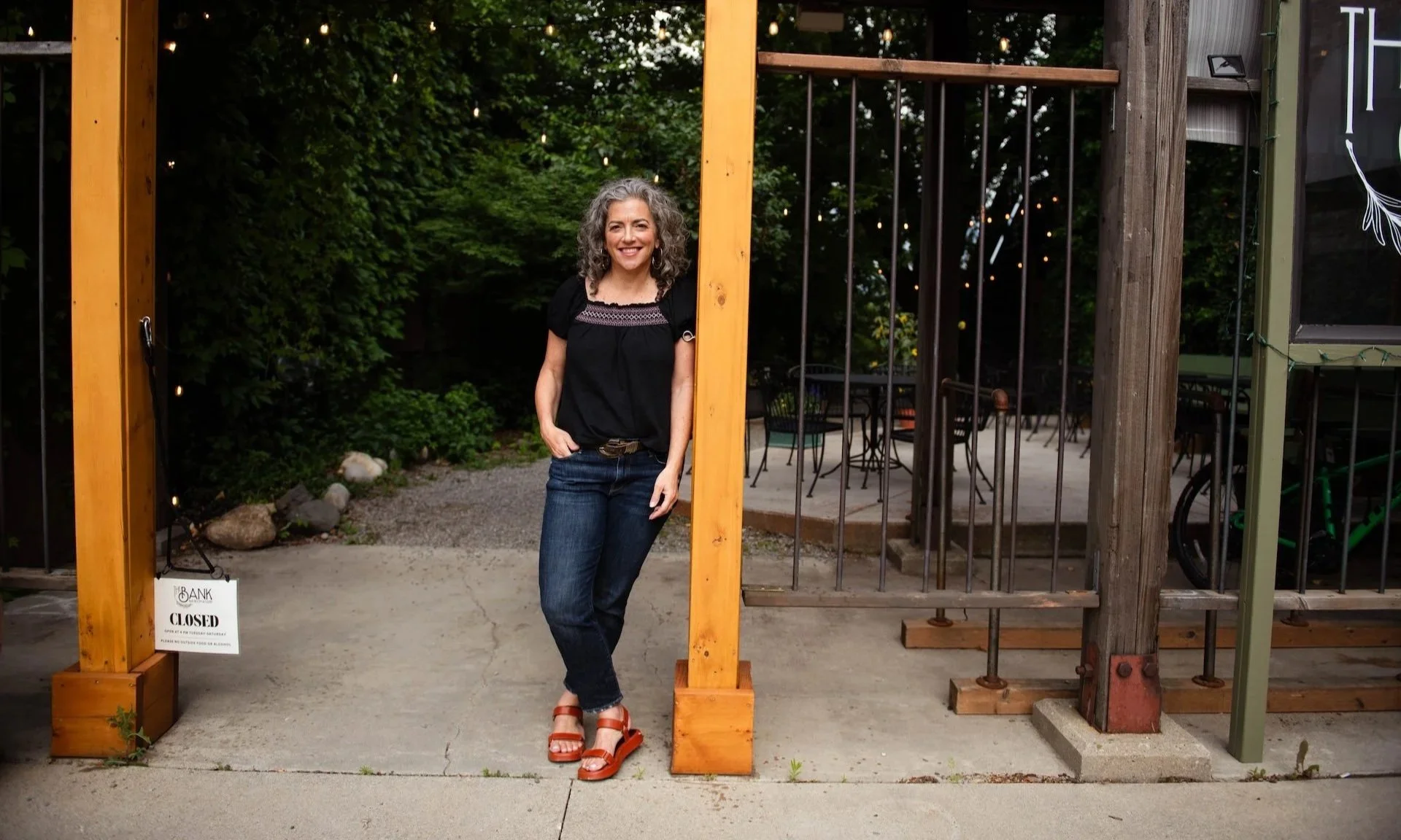Woman standing in front of a closed restaurant entrance with a 'Closed' sign, outdoor patio with tables, chairs, and trees in the background.