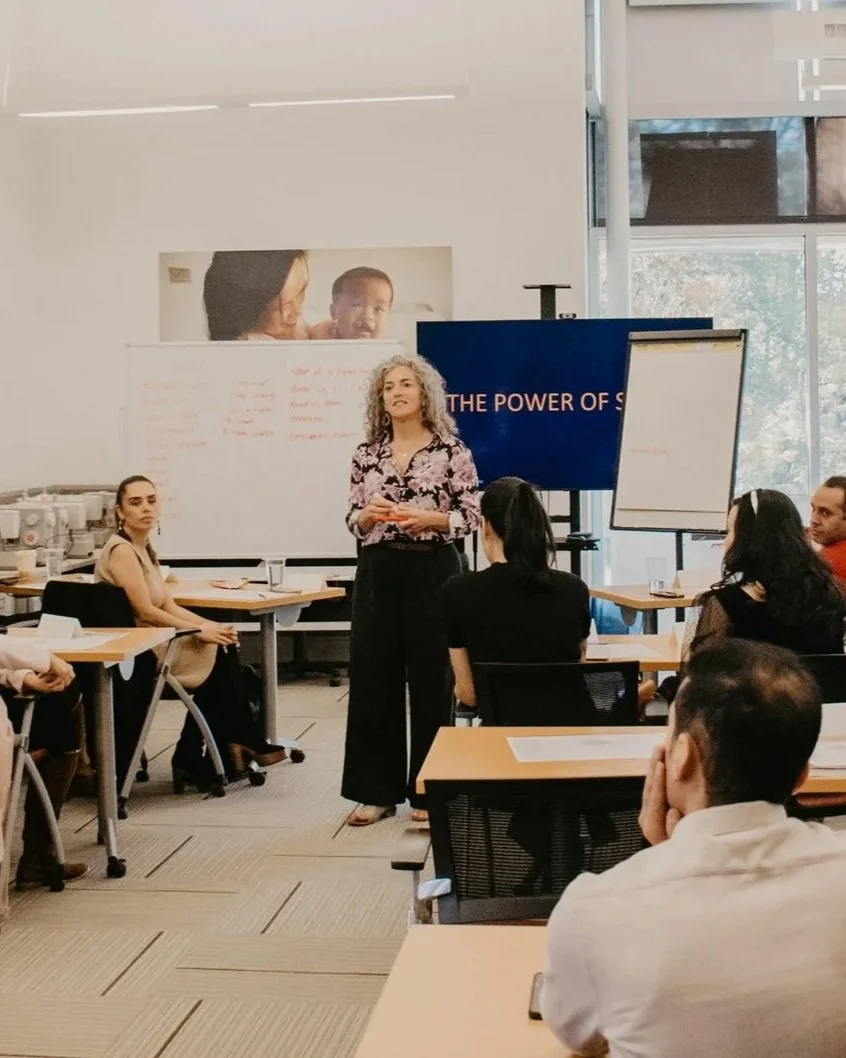 A woman giving a presentation to a group of people in a conference room with a whiteboard, a large screen displaying 'The Power of S', and a flip chart. There is a window showing trees outside.