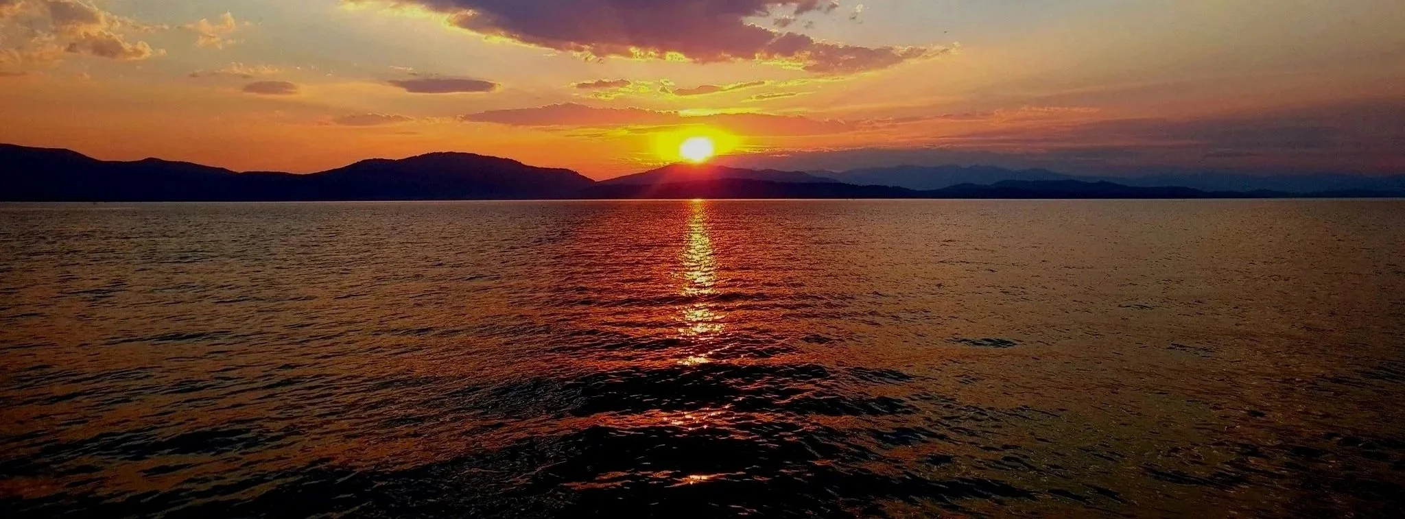 A sunset over a large body of water with silhouettes of distant mountains on the horizon. The sky has vibrant colors including orange, yellow, and purple, reflected on the rippling water surface.