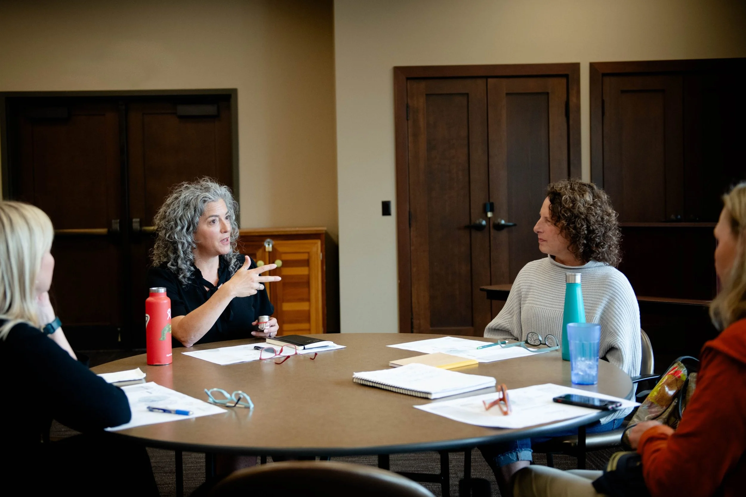 A woman with curly gray hair is speaking during a meeting with four other women at a conference table. The table has notebooks, pens, water bottles, and glasses. The room has wooden doors and beige walls.