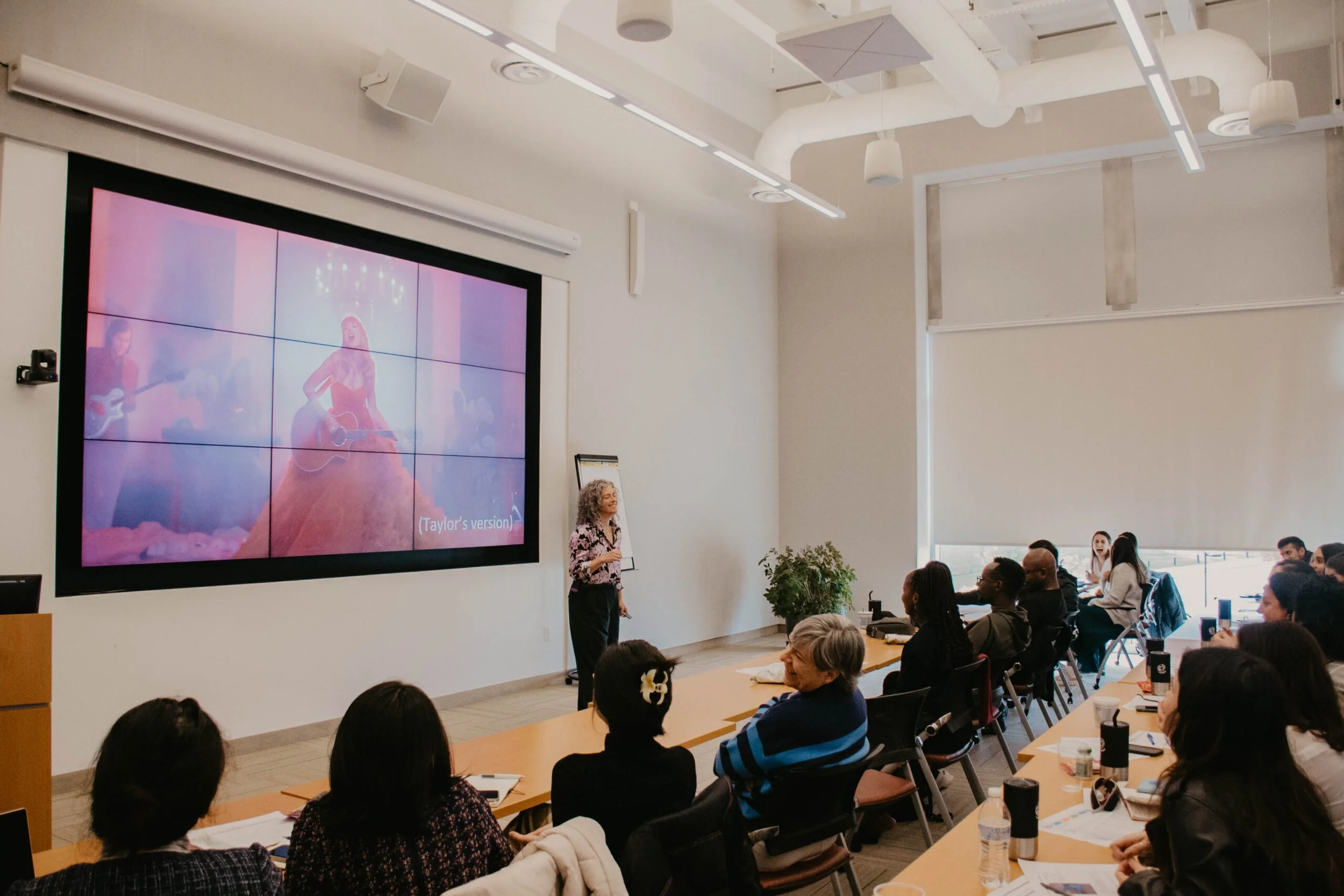 A woman giving a presentation to a seated audience in a bright, modern conference room with a large screen displaying an image of a woman with a guitar.