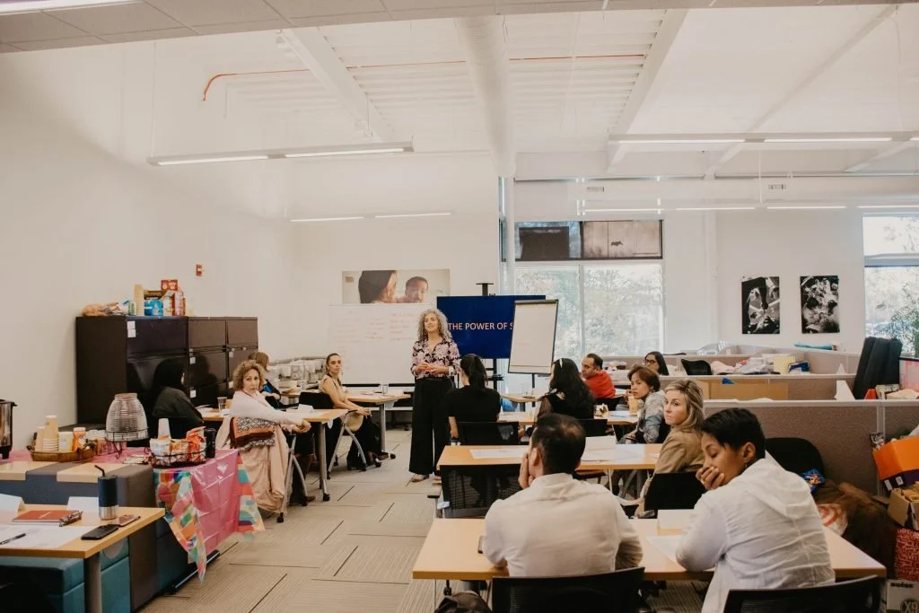 A diverse group of adults attending a presentation or workshop in a bright, modern office or conference room with large windows and minimal decor.
