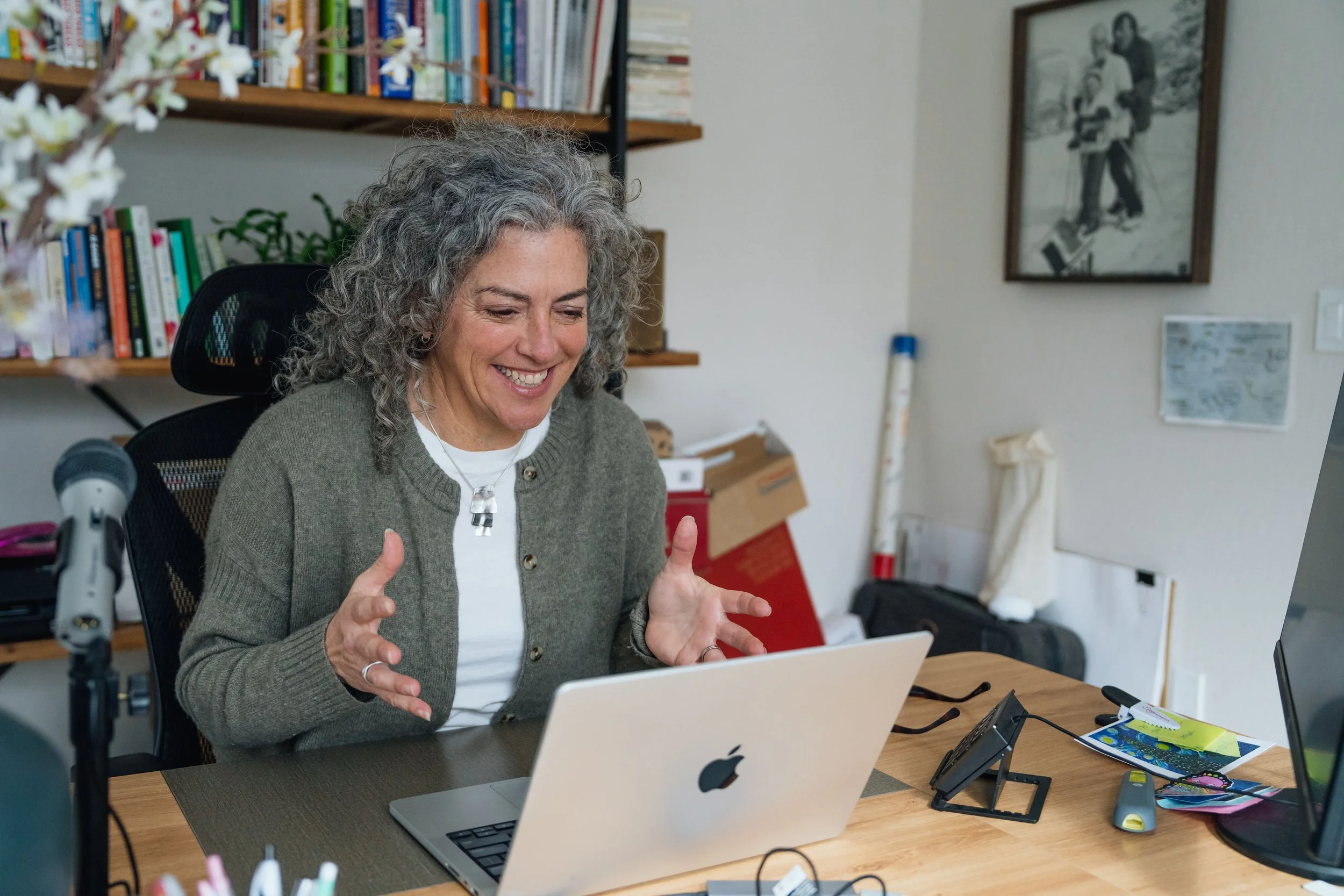 A woman with curly gray hair smiling and gesturing with her hands while sitting at a desk with a laptop, in an office with bookshelves and framed pictures on the wall.