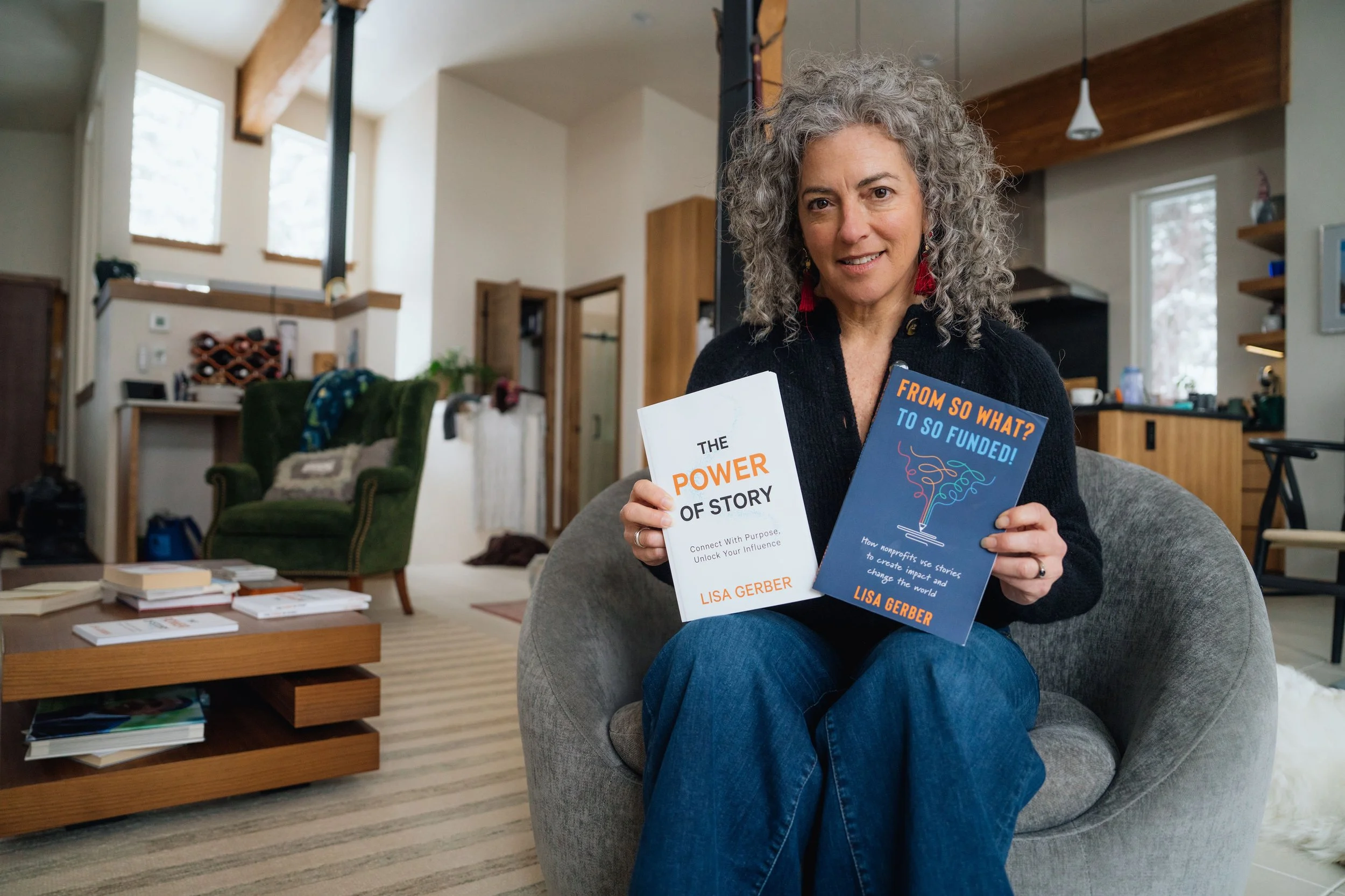 A woman with curly gray hair sitting on a gray chair in a living room, holding two books titled 'The Power of Story' and 'From So What? To So Funded!'