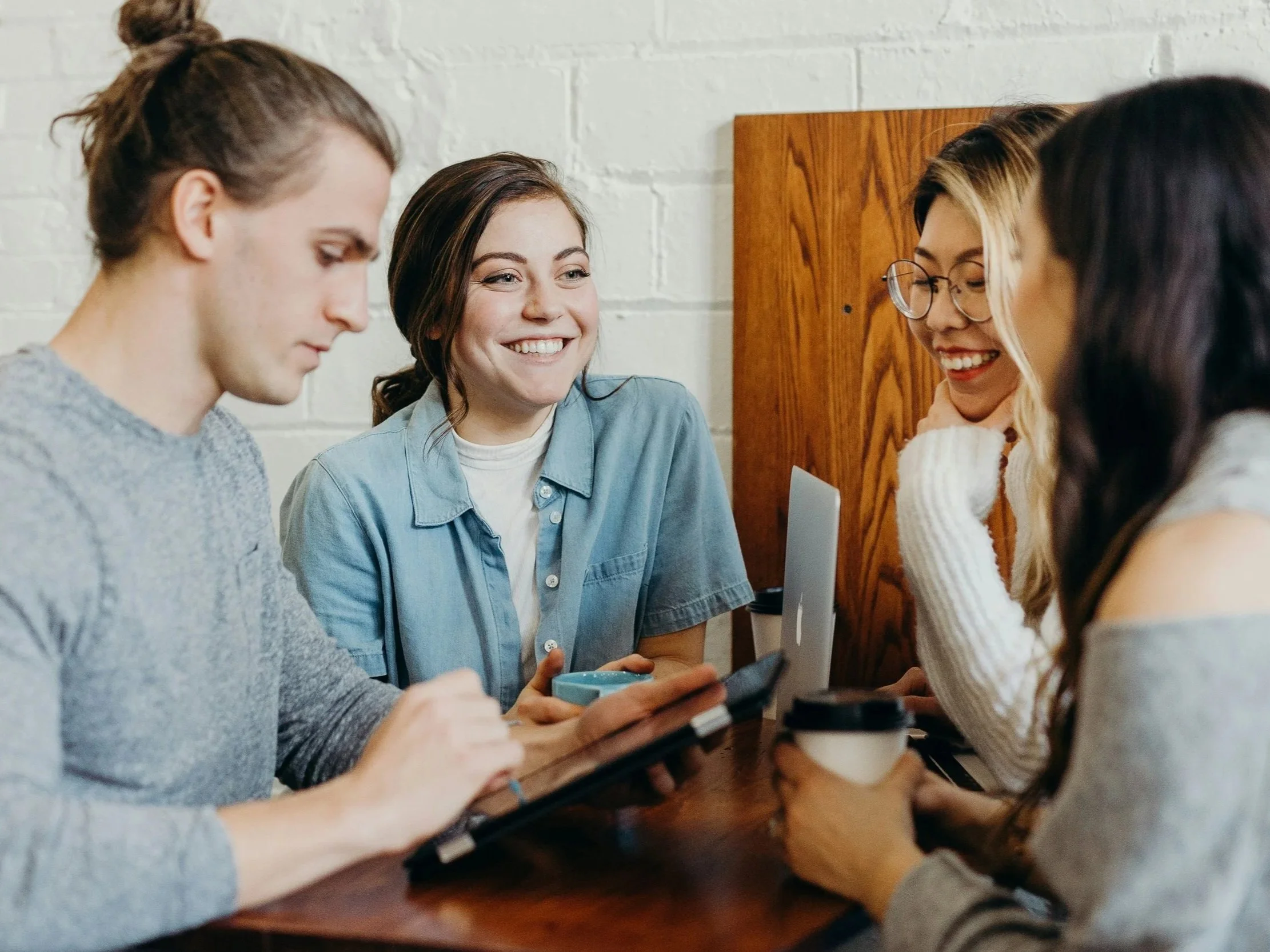 Group of young women gathered around a wooden table, smiling and looking at a smartphone, in a casual indoor setting with white brick walls.